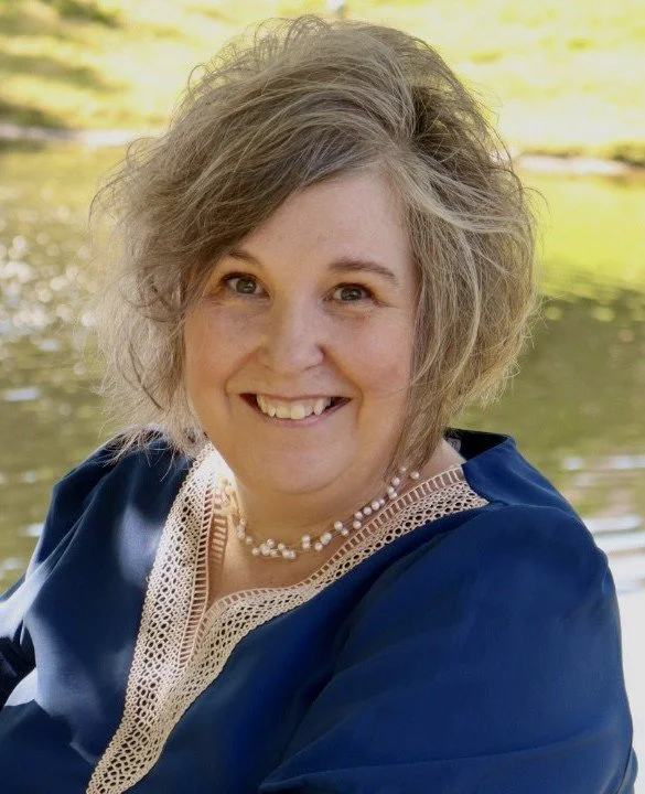A woman with curly, shoulder-length gray hair smiling outdoors near a body of water, wearing a navy blue dress with lace detail and a pearl necklace.
