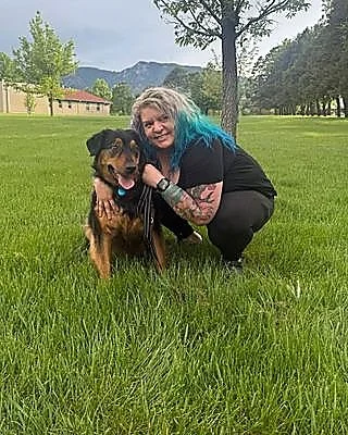 A woman with blue hair posing with her black and brown dog in a grassy park beneath a tree with mountains in the background.