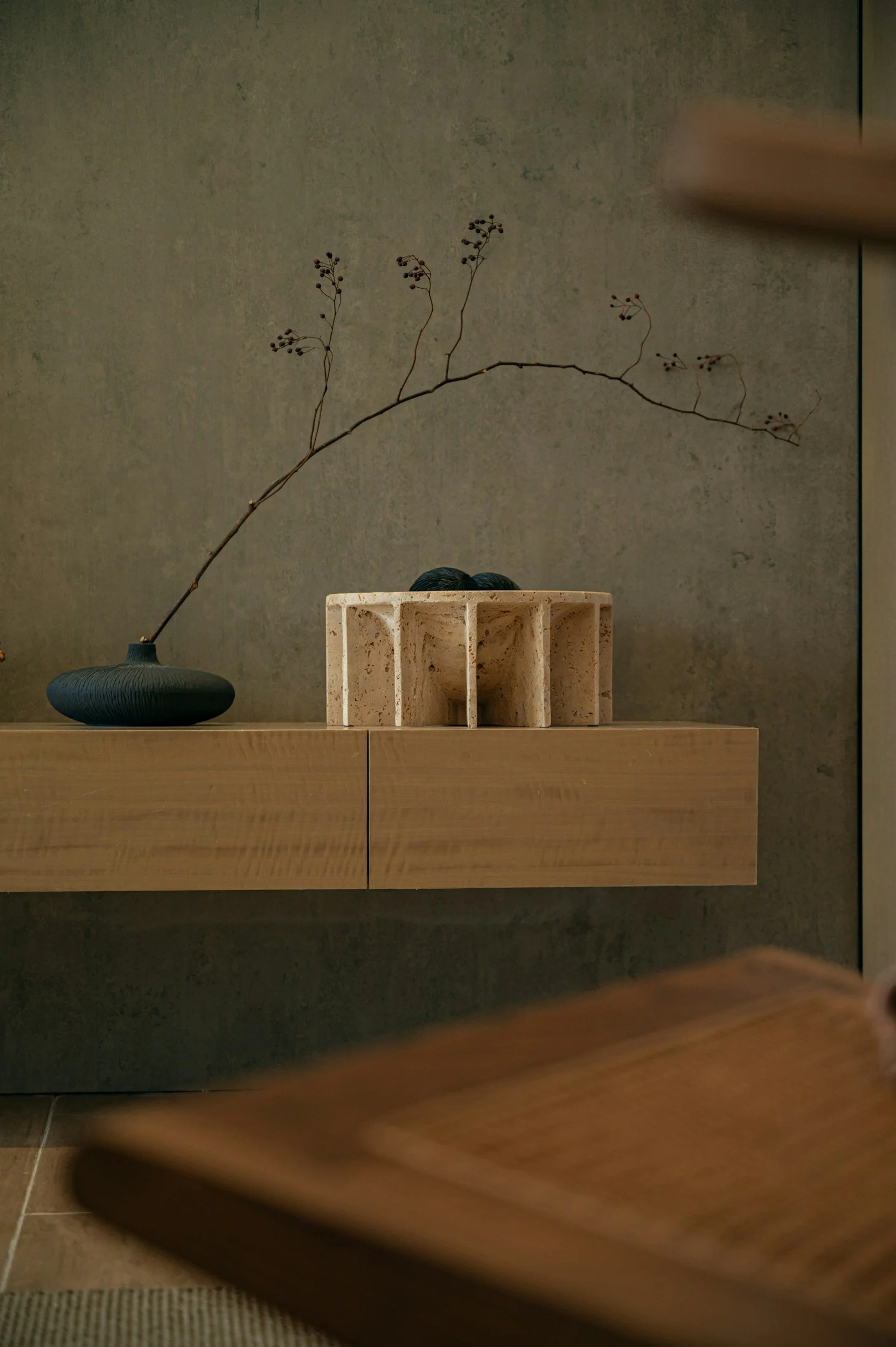 A minimalist wooden shelf with a black vase holding a long, thin branch with small berries, and a beige textured ceramic container. Part of a wooden chair is visible in the foreground, set against a plain wall.