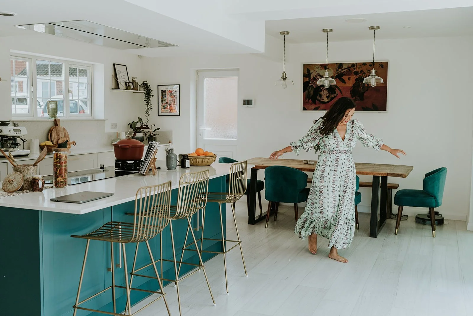 A woman in a patterned dress walking barefoot in a bright, modern kitchen and dining area with white walls and light wooden floors. There are teal and gold bar stools at the kitchen island, a table with teal chairs, and various decorations on the walls and counters.
