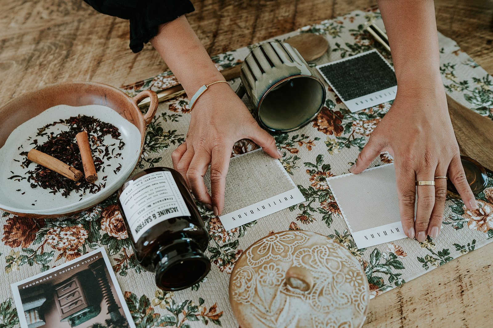 Person arranging fabric sample swatches on a floral tablecloth surrounded by tea leaves, a jar, a mug, a sugar bowl, a tea infuser, and a photo of a drawer.