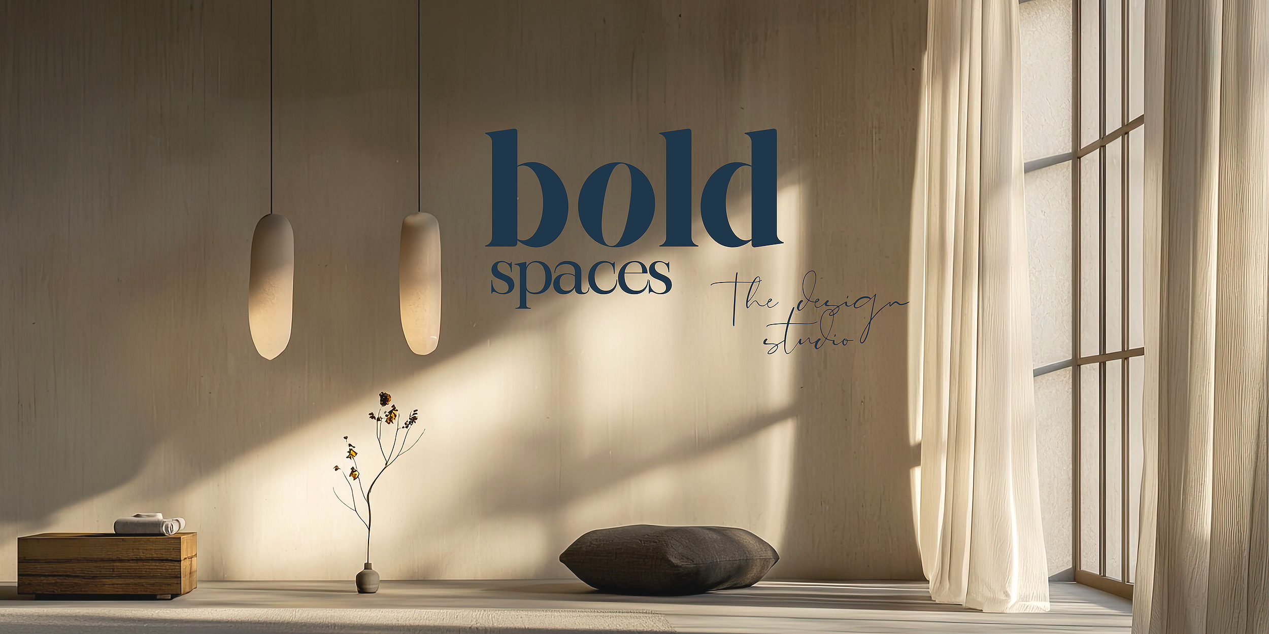 Interior of a minimalist design studio with two hanging pendant lights, a small wooden table with rolled towels, a decorative branch in a vase, a dark cushion, and sheer curtains beside large windows with sunlight streaming in.