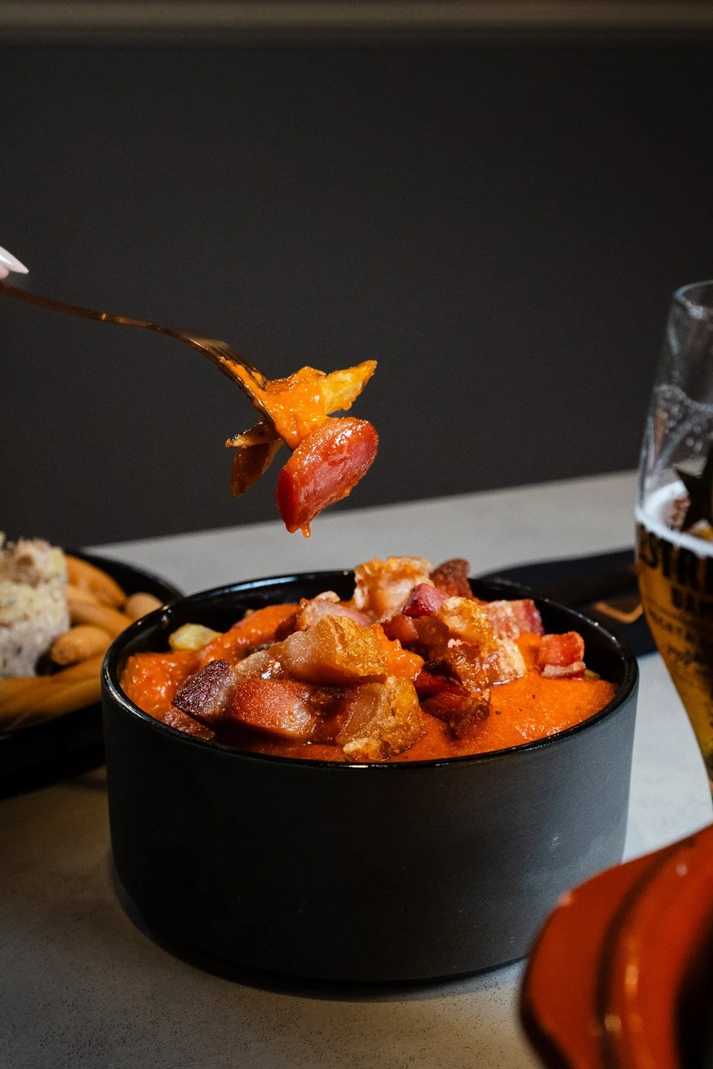 Una porción de comida en un cuenco negro, con torreznos de Soria y salsa, con un tenedor levantando un bocado, acompañado de una bebida y aperitivos en platos a su lado.