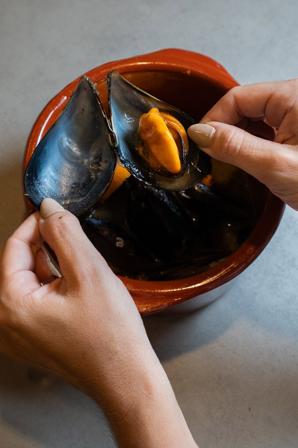 Manos sosteniendo una concha de mejillón con carne de mejillón dentro. En un recipiente grande de cerámica marrón.