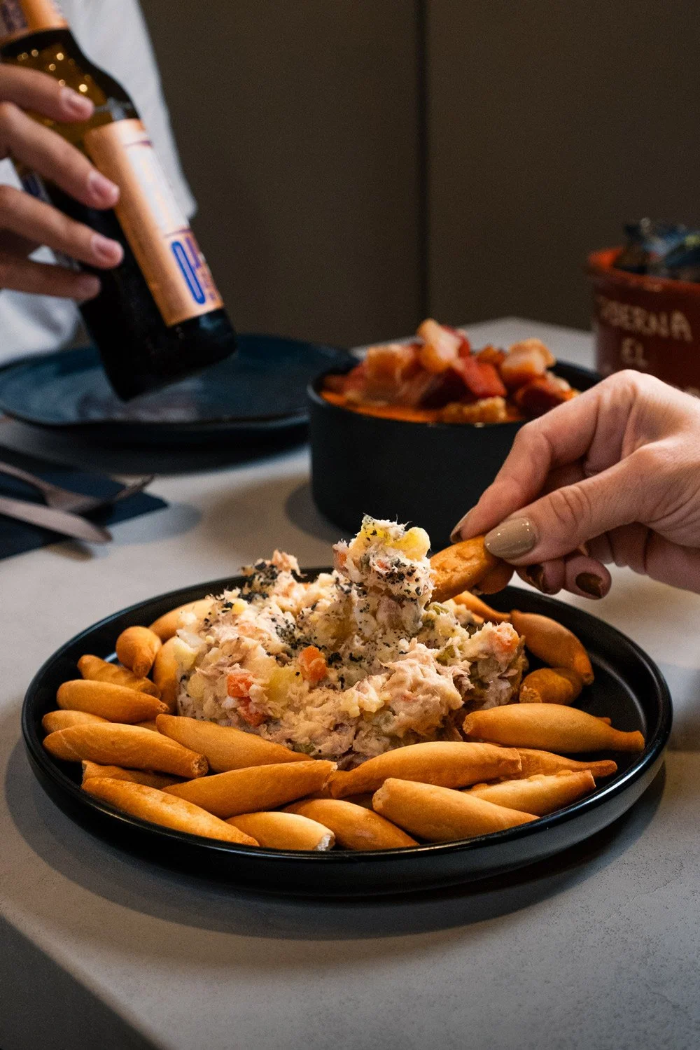 Persona comiendo ensaladilla con galletas saladas en un plato negro.