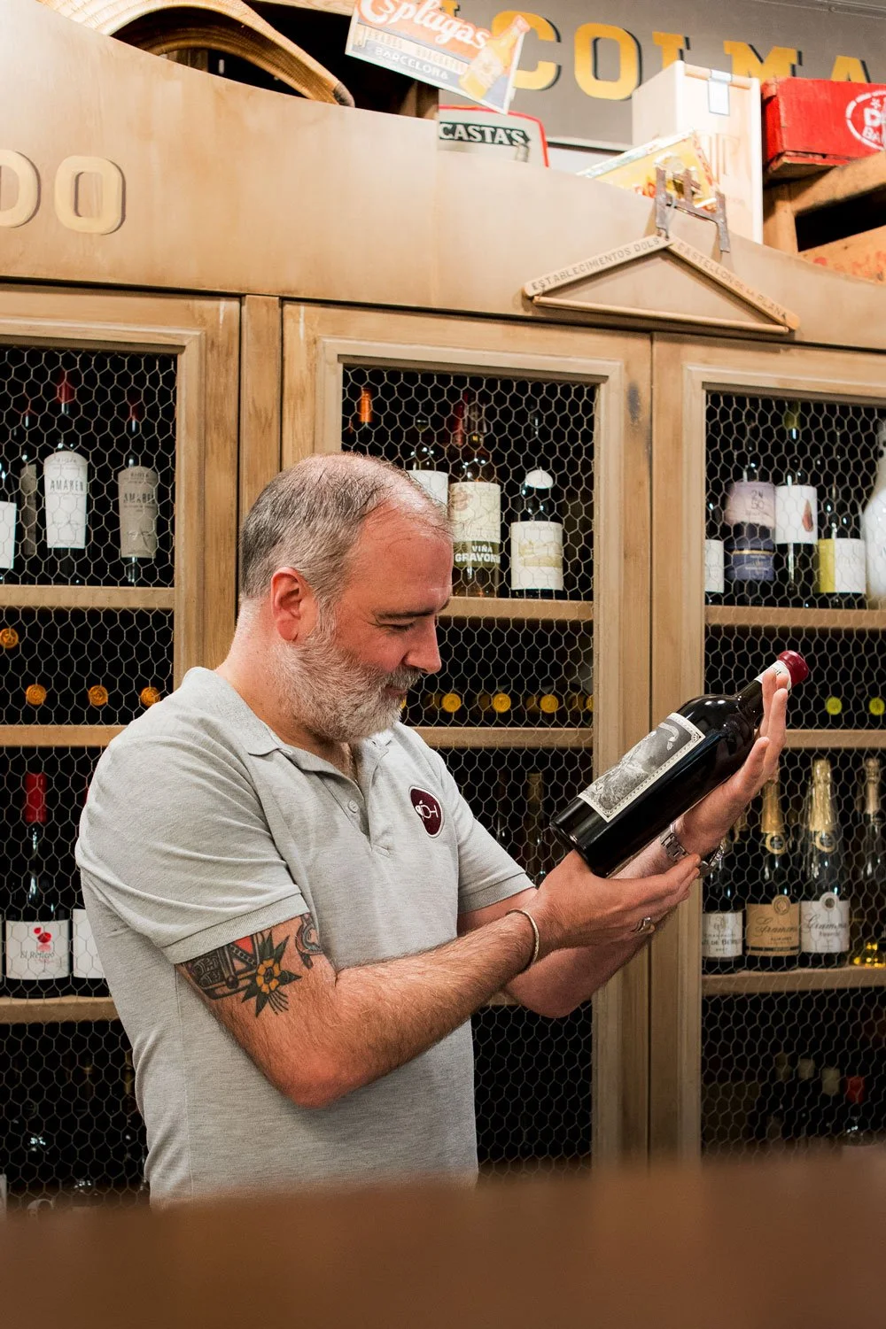 Hombre con barba y tatuajes sosteniendo una botella de vino en una tienda de vinos, con estanterías llenas de botellas de vino y decoraciones de madera en el fondo.