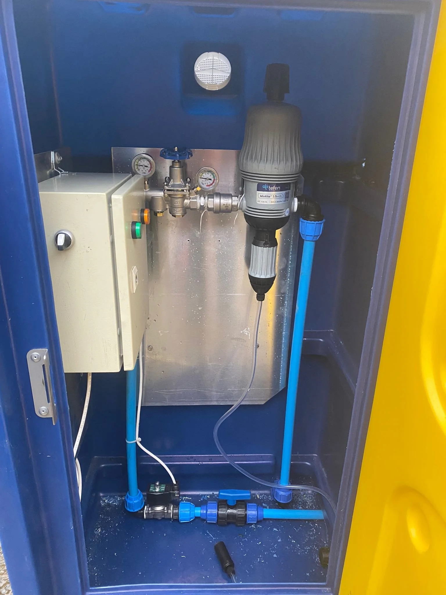 A water treatment or filtration system inside a blue and yellow cabinet, featuring a control box, gauges, a pump, blue piping, and valves.