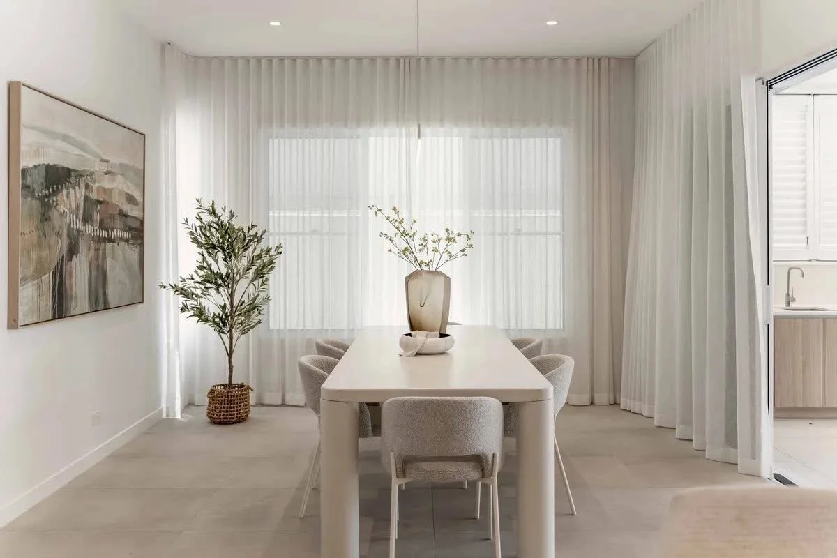 Minimalist dining room with a white table, six light-colored chairs, a tall vase with flowers, a framed abstract painting, a potted plant, and large windows with white curtains.