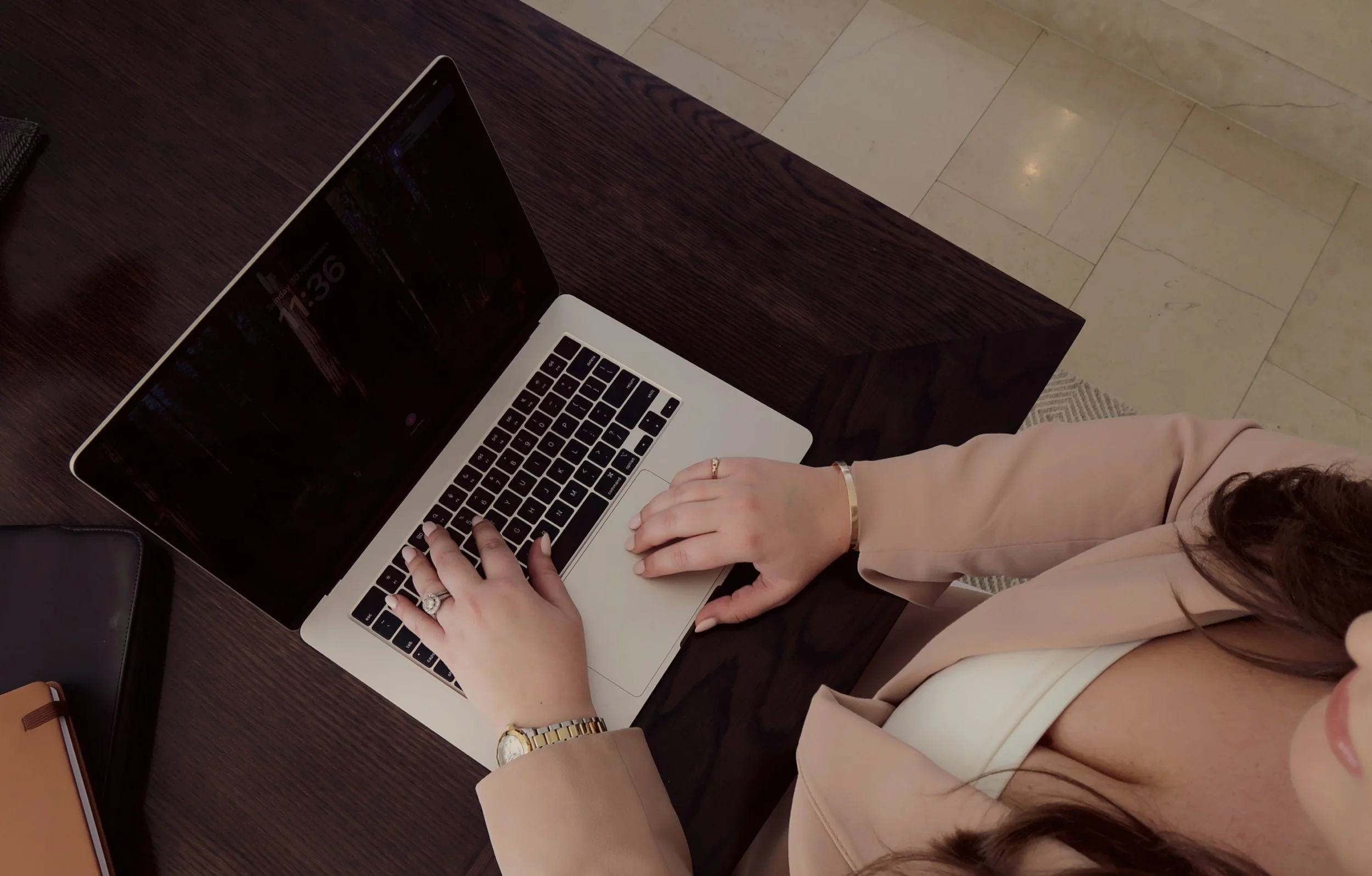 A woman using a MacBook laptop on a wooden desk, wearing a beige blazer and jewelry.