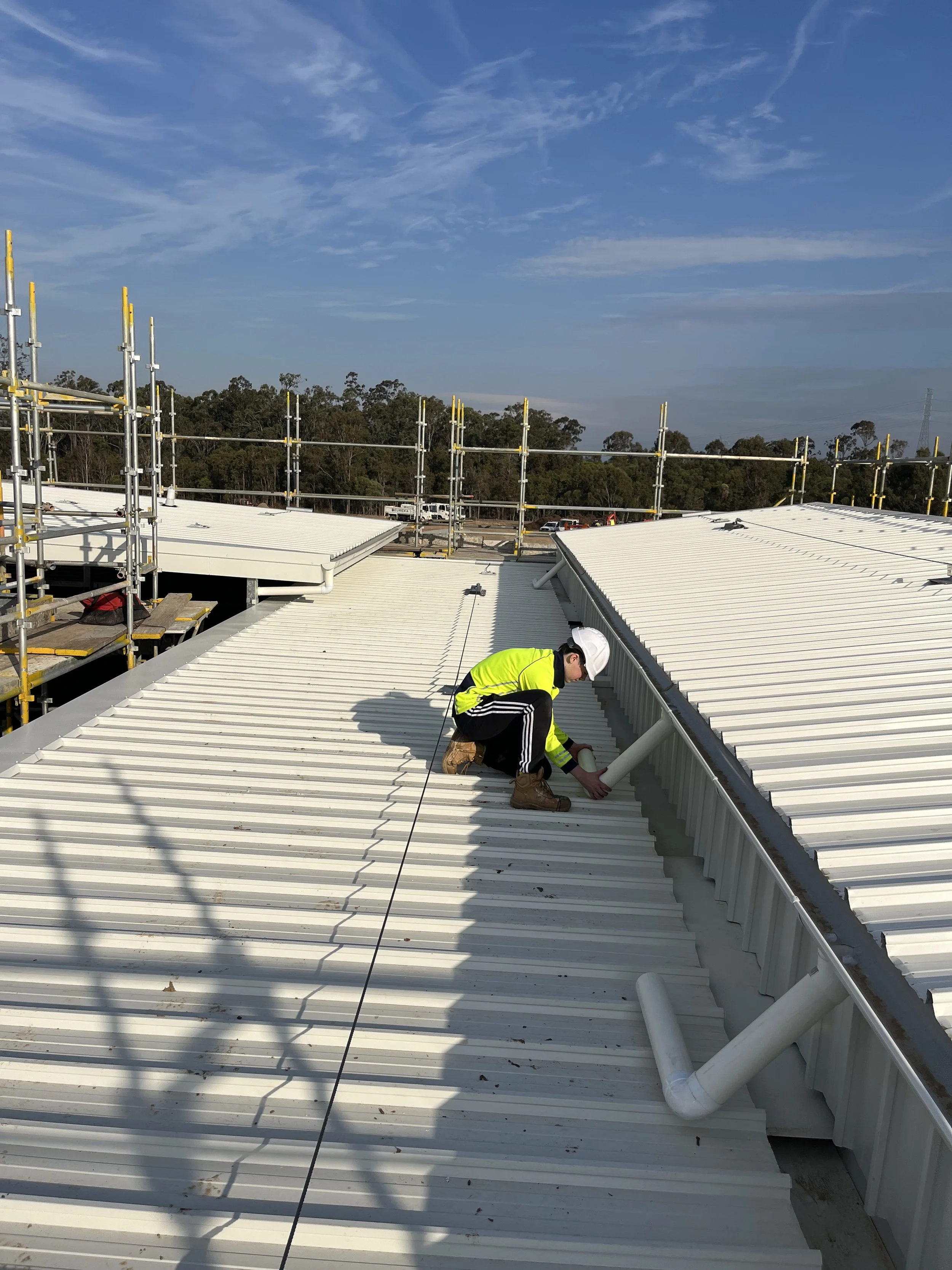 Worker wearing a yellow safety vest and white helmet crouching on a white metal roof, installing or inspecting a white vent pipe, with scaffolding and a forested area in the background under a blue sky with scattered clouds.