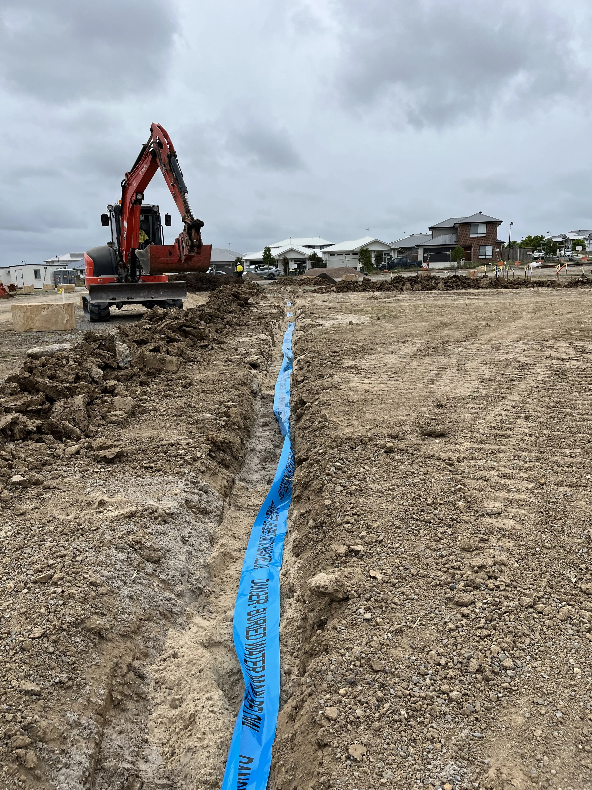 Construction site with excavator digging a trench, blue utility marking tape in the ground, and nearby residential houses under a cloudy sky.