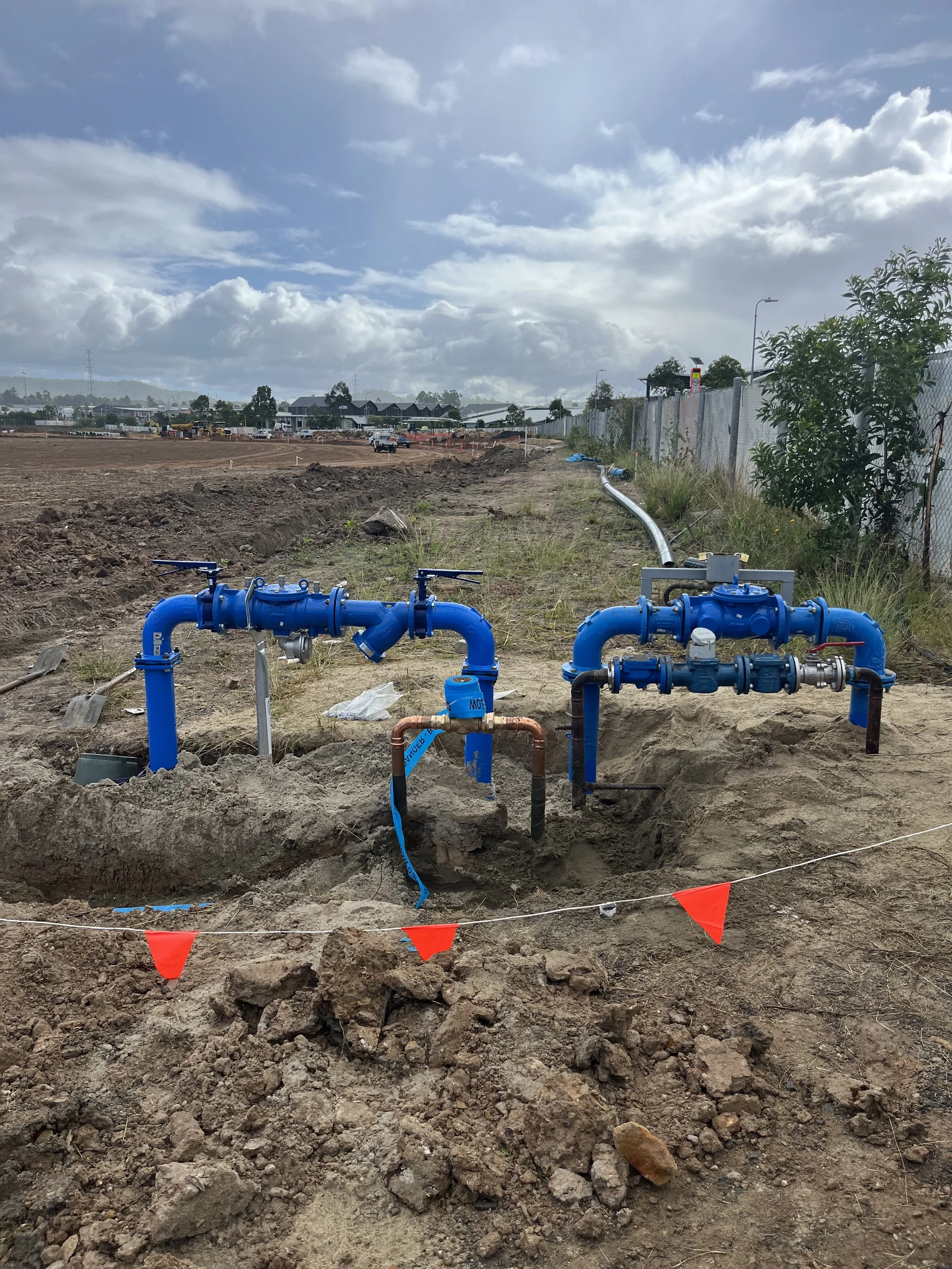 Construction site with blue water pipes installed in the ground, orange flags marking the area, and a dirt landscape with cloudy sky in the background.