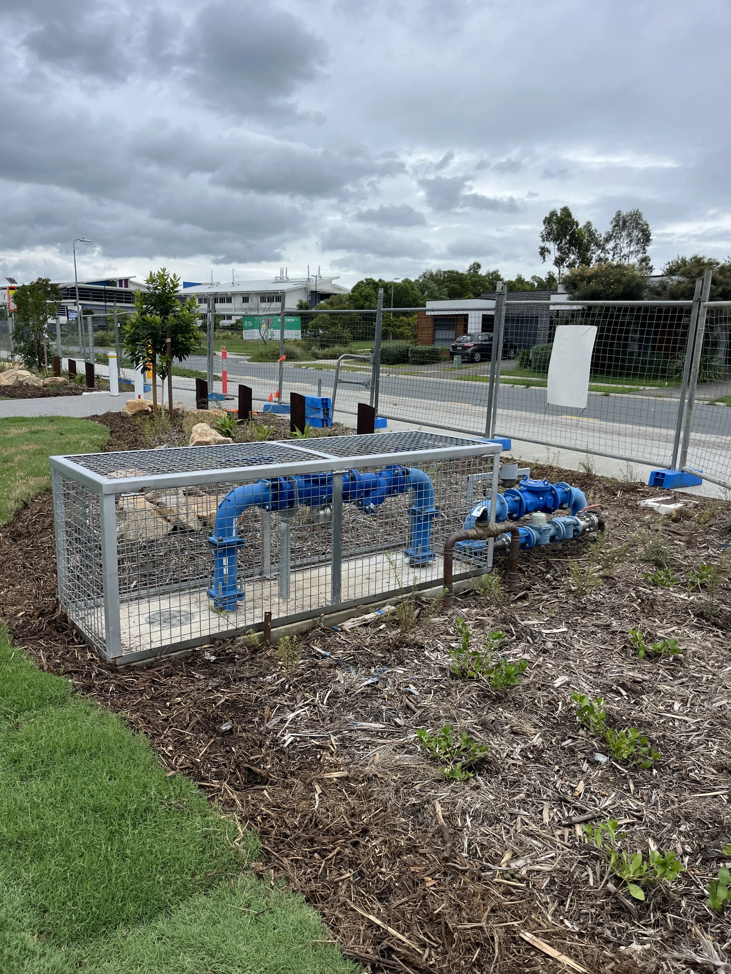 Construction site with blue piping, a fenced area, and a landscaped background with trees and modern buildings under a cloudy sky.