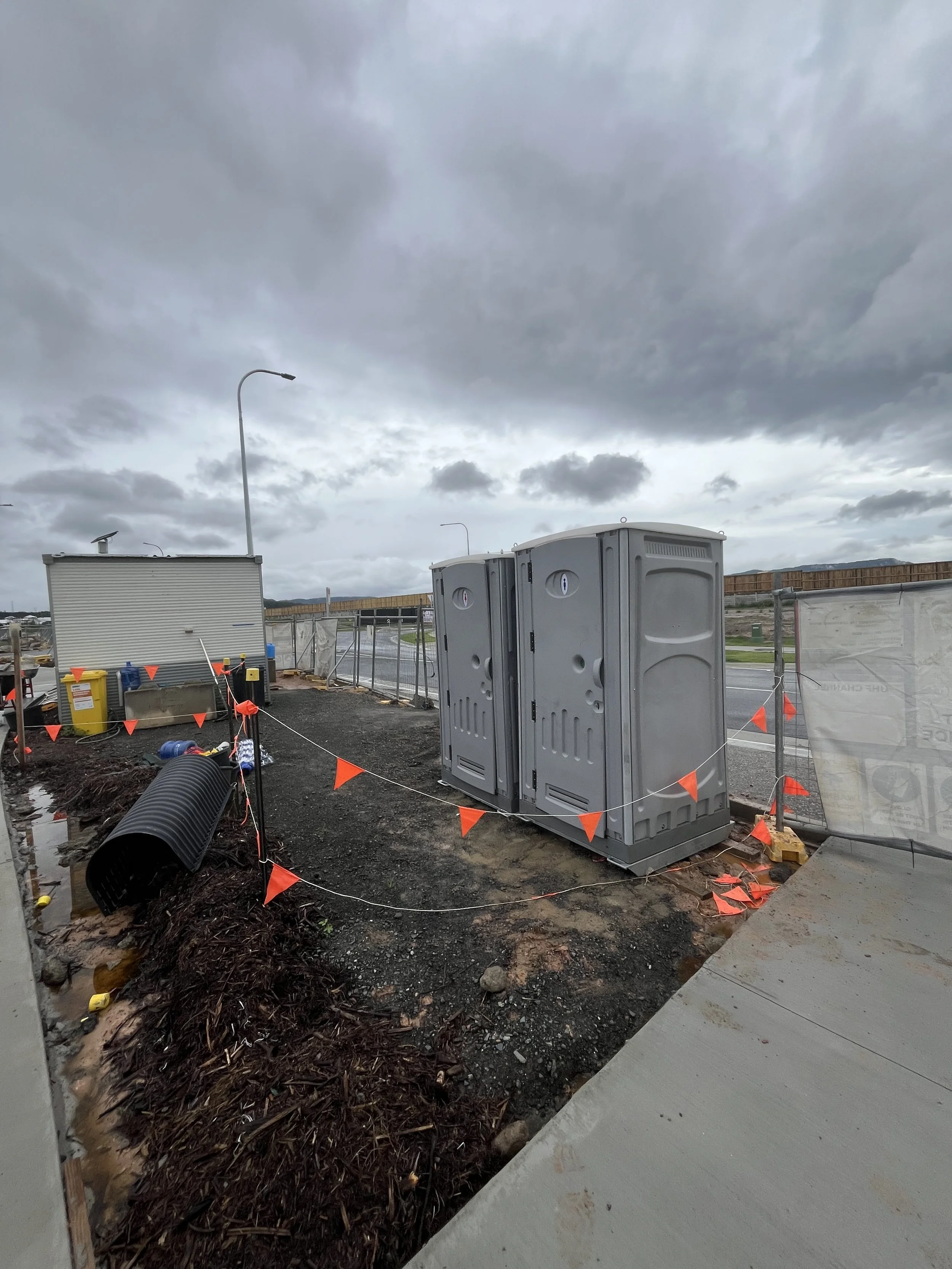 Construction site with portable toilets, an electrical box, and a dirt pathway under cloudy skies.