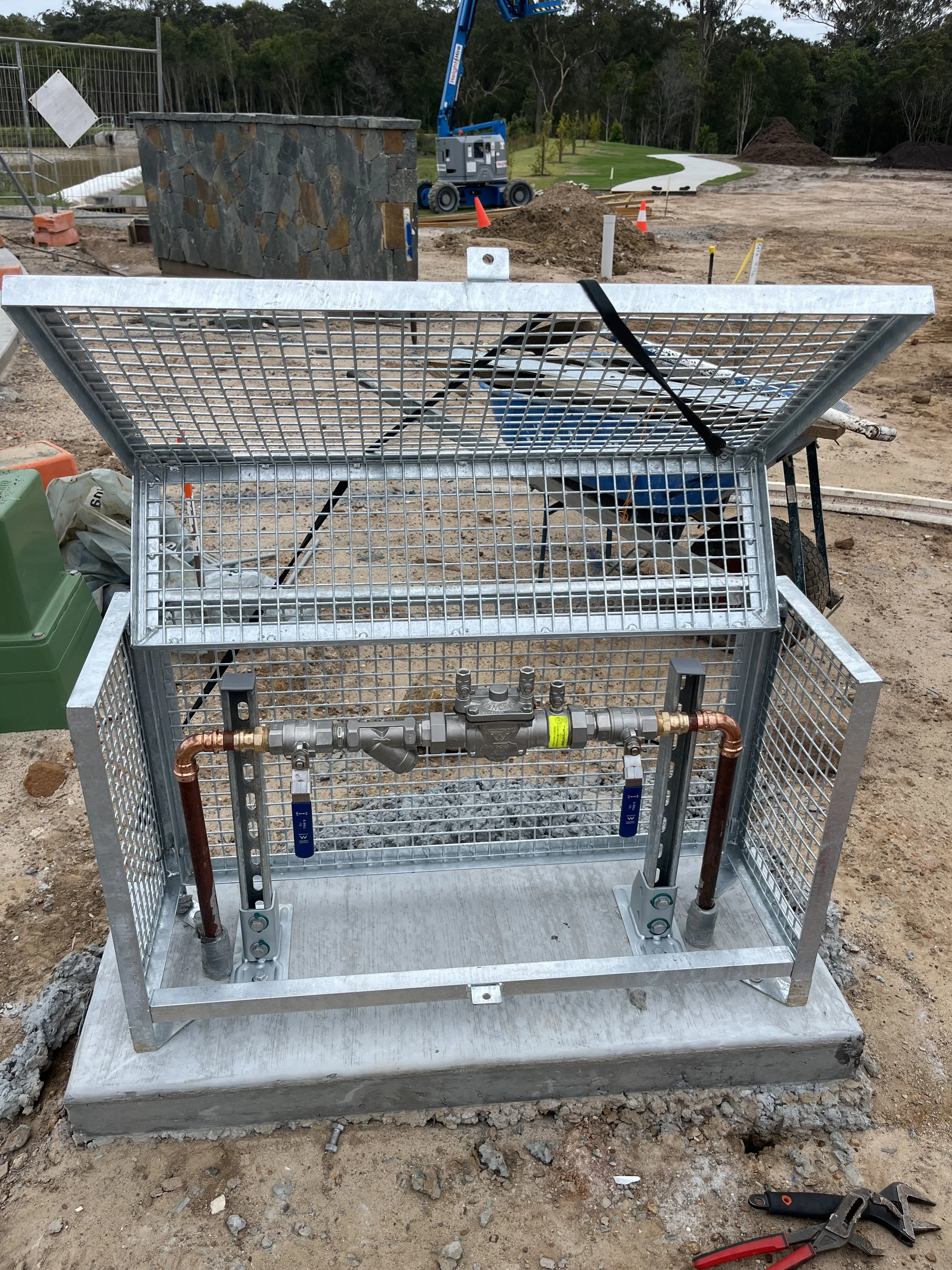 Construction site with a metal utility box with plumbing pipes and gauges at the front, outside in a cleared area with piles of dirt, orange safety cones, a gravel pathway, trees in the background, and construction equipment.