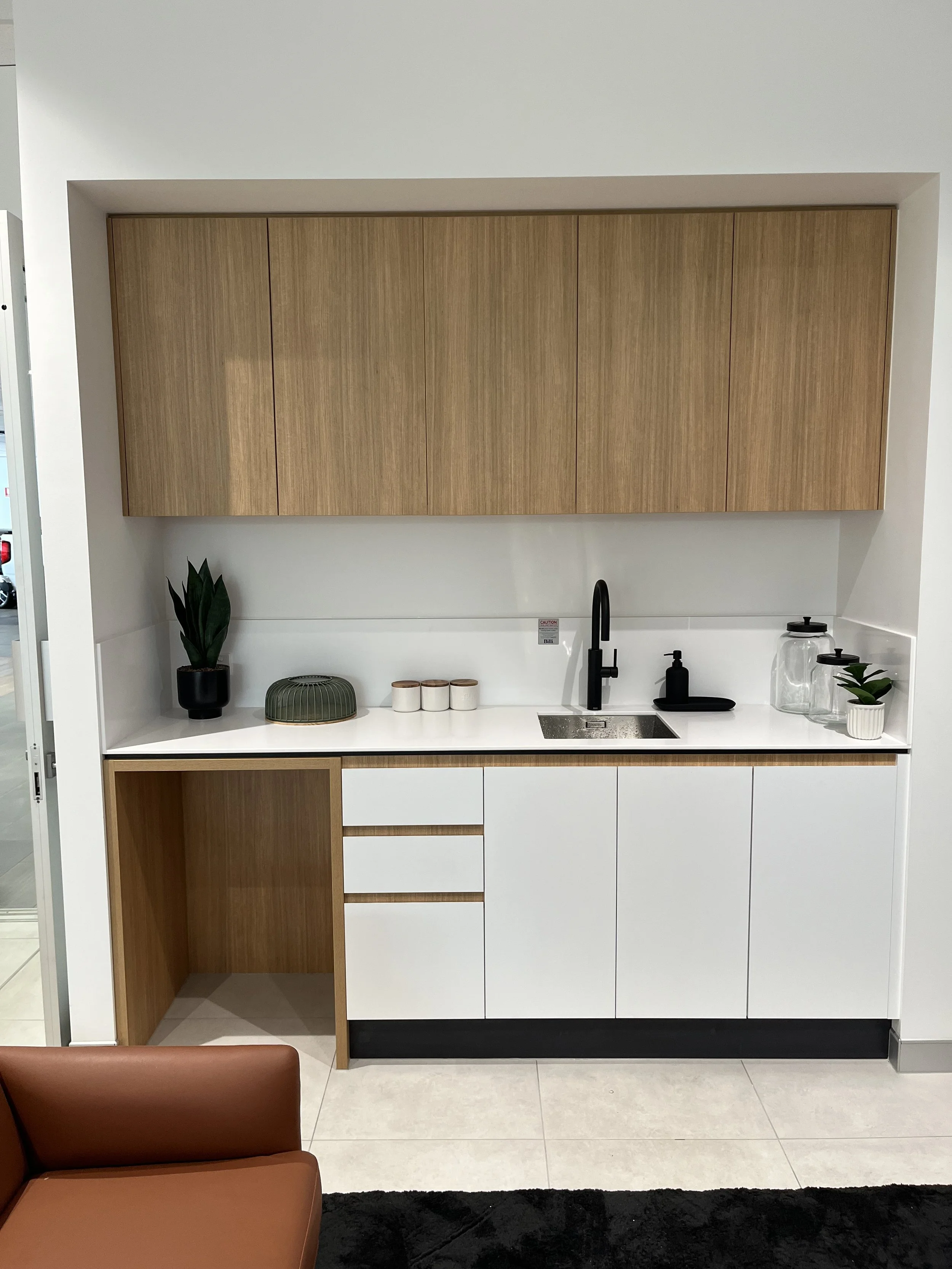 Modern kitchen area with wooden upper cabinets, white lower cabinets, a black faucet, and a small potted plant on the right side of the counter. There are some decorative jars and a plant on the counter, and a brown chair in the foreground. The floor
