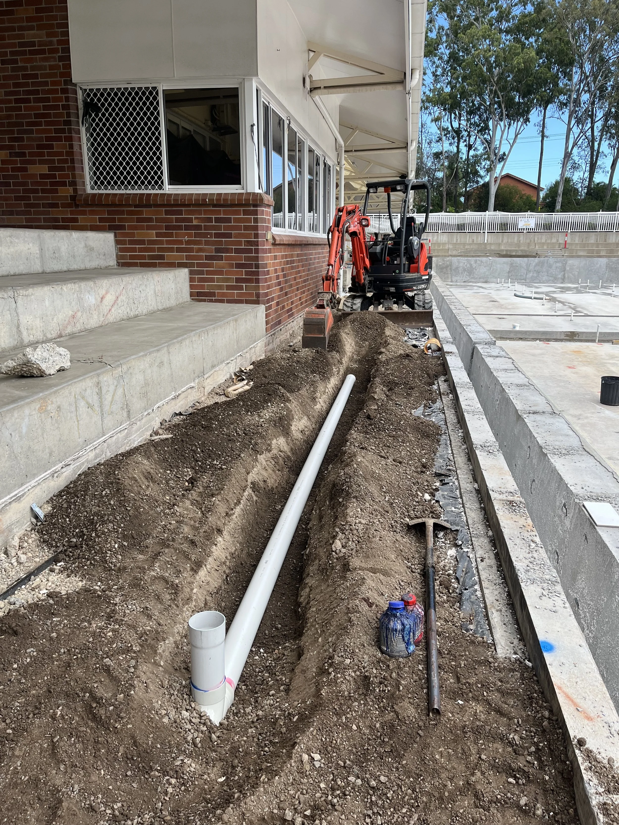 Construction site with exposed soil, pipes, and construction tools; building exterior with brick wall and large windows; ongoing plumbing or drainage work.