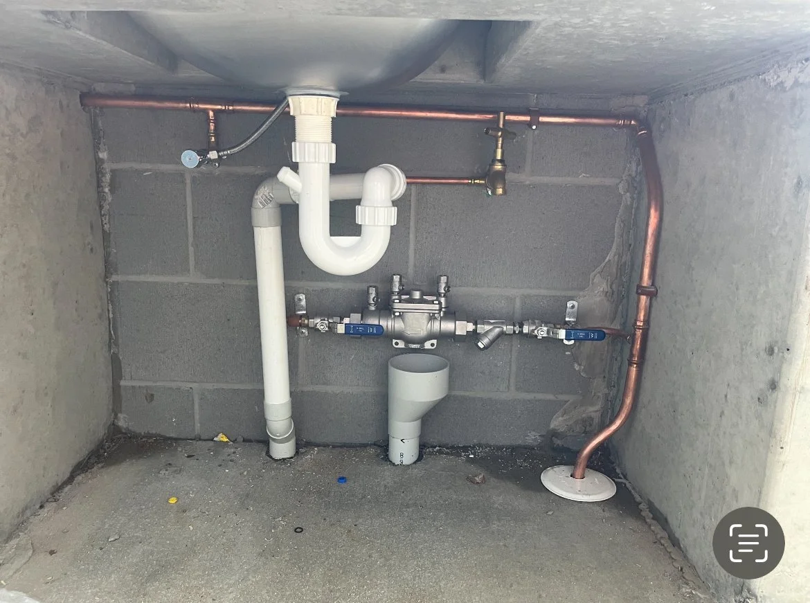 Plumbing pipes and valves installed in a wall cavity, including white PVC pipes, copper pipes, a trap, and shut-off valves.