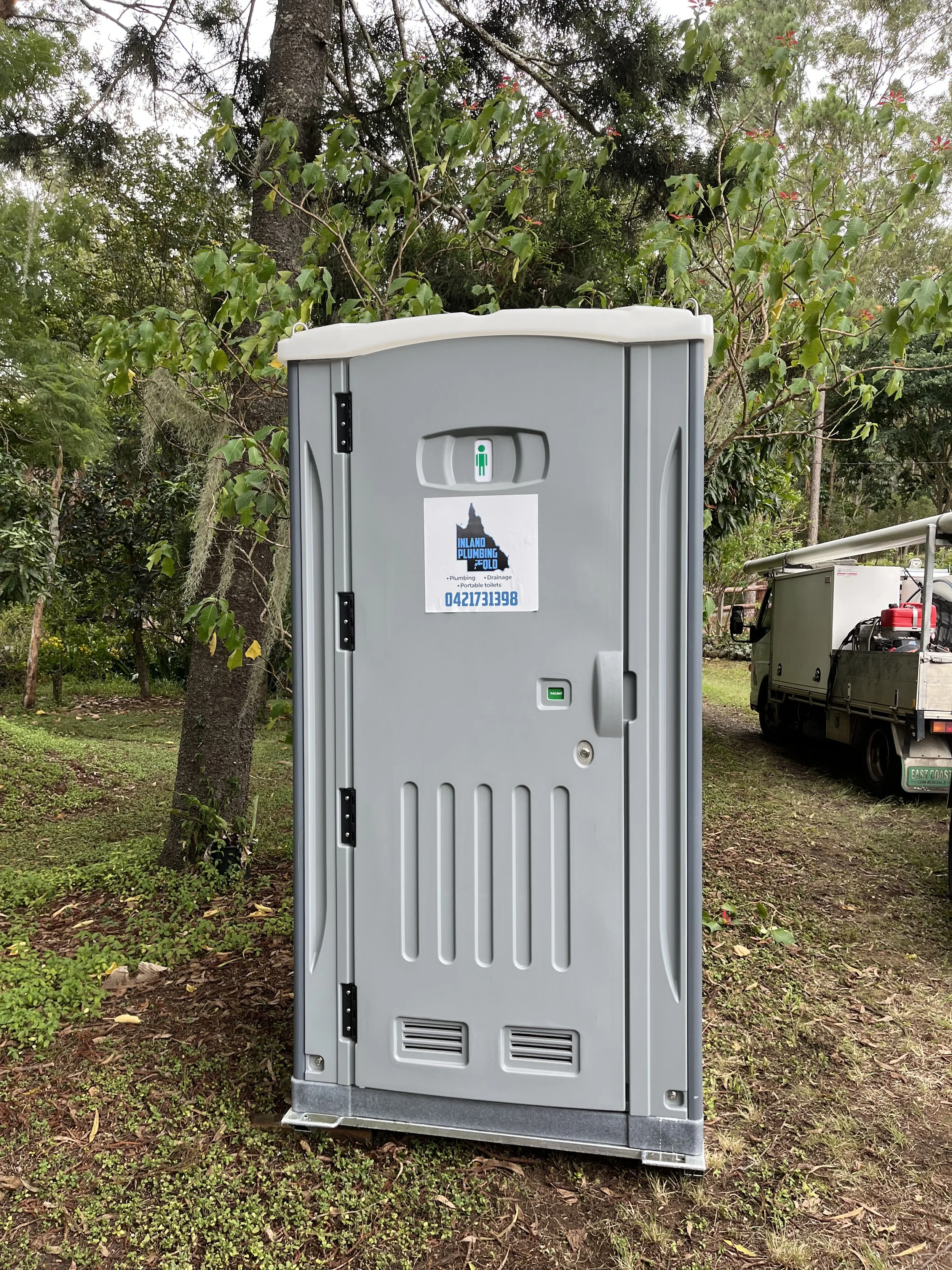A portable toilet unit in a natural outdoor setting with trees and grass around, and a utility vehicle nearby.