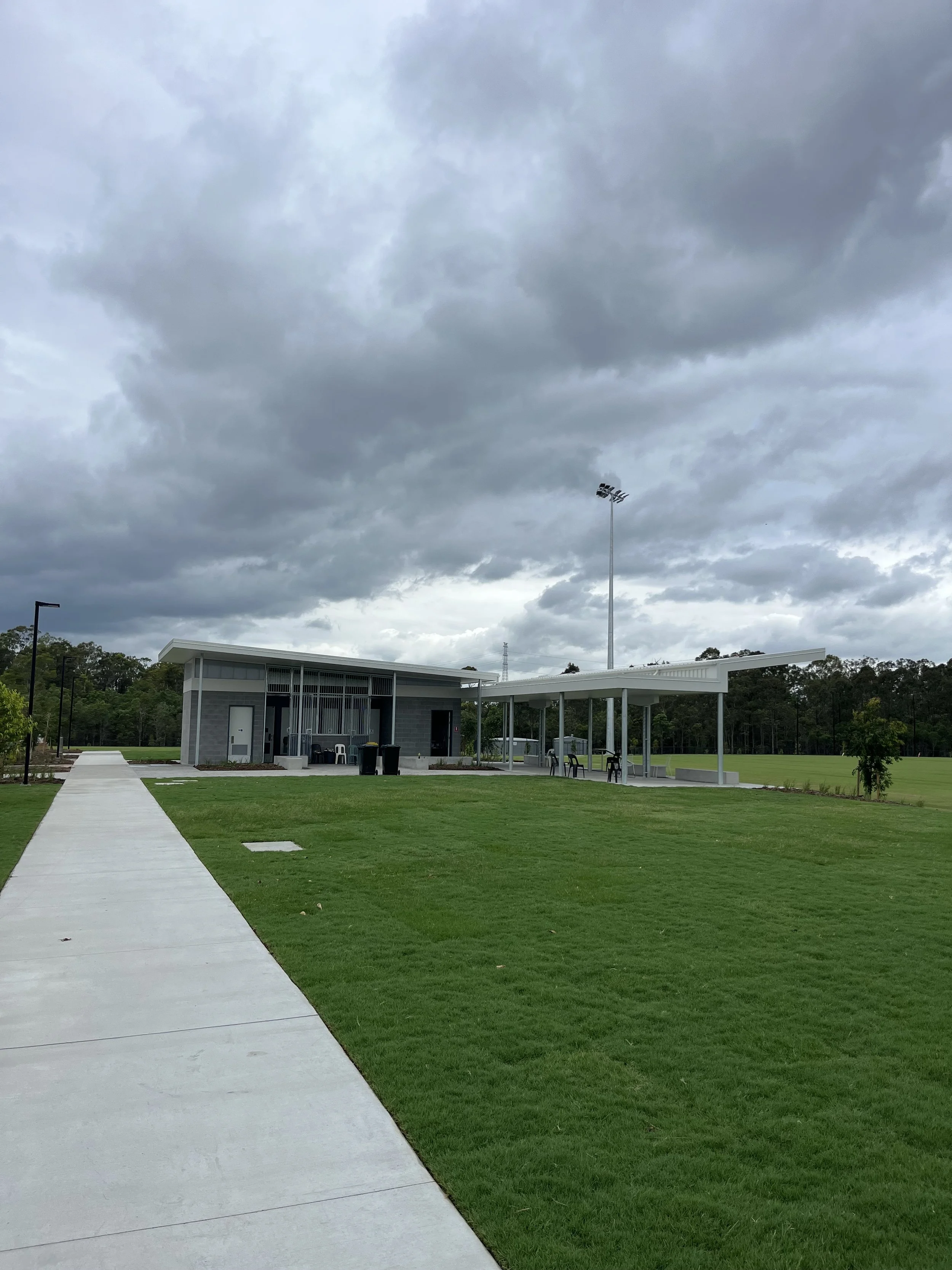 A park with a concrete pathway, green grass, and a small building with a covered area, set under dark cloudy skies.