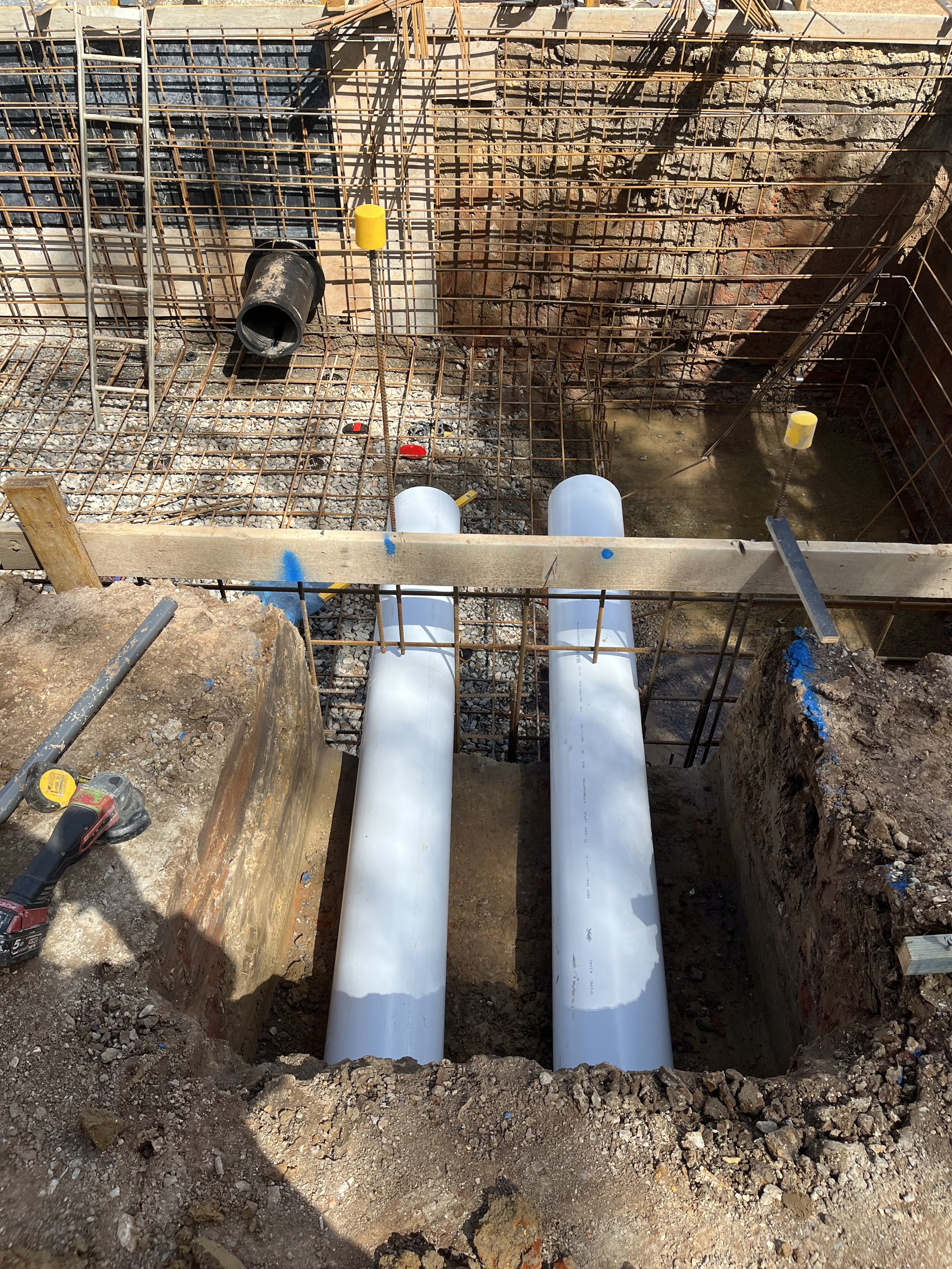 Construction site with two large white plumbing pipes running horizontally through a deep excavated trench, metal rebar grid surrounding the pipes, and construction tools and materials scattered around.