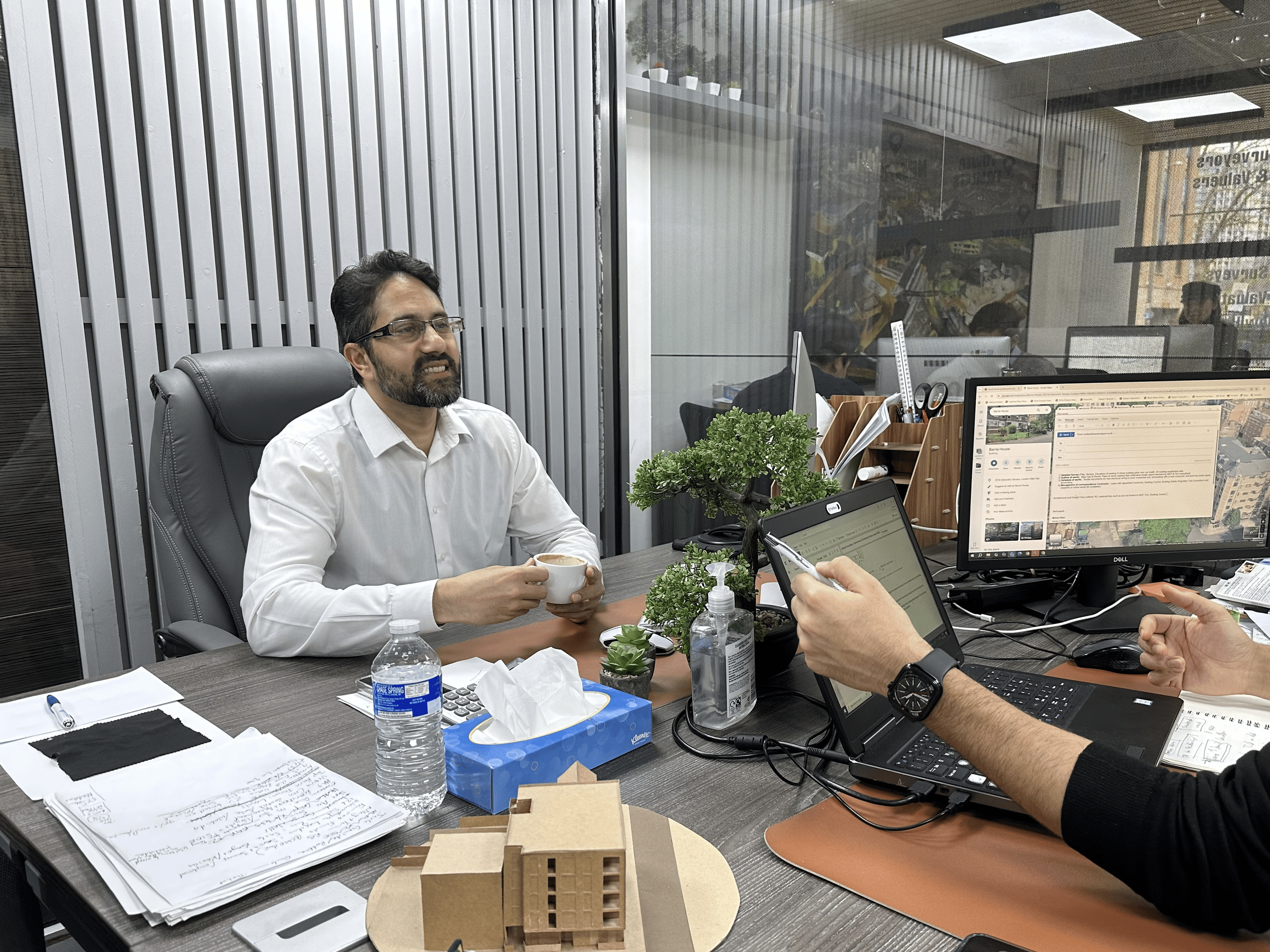 A man with glasses and a beard wearing a white shirt sitting at a desk with a coffee cup, talking to someone with a tablet, in an office with multiple computer monitors and office supplies.