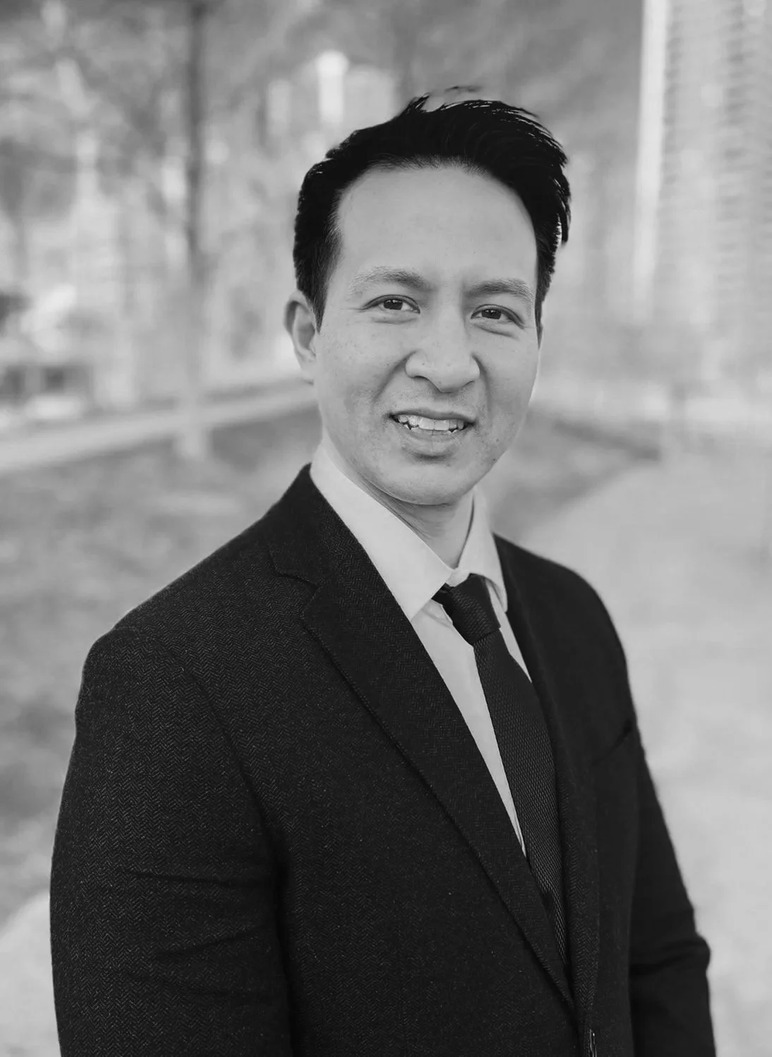 Black and white portrait of a man in a suit smiling outdoors with a blurred background of trees and a brick building.