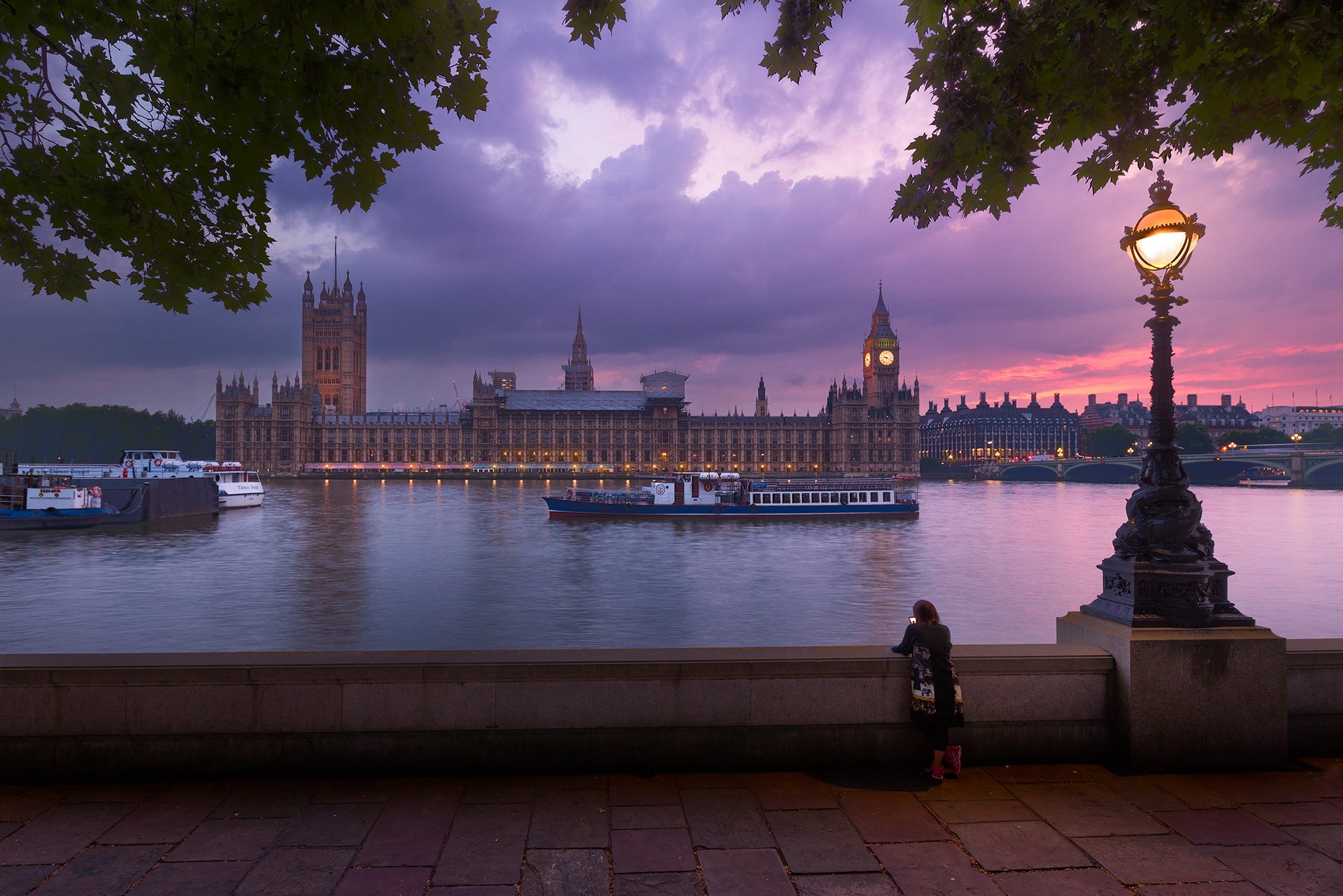 A young girl standing by a riverside wall in London, watching a boat pass on the River Thames with the Houses of Parliament and Big Ben in the background during sunset, framed by trees and a vintage street lamp.