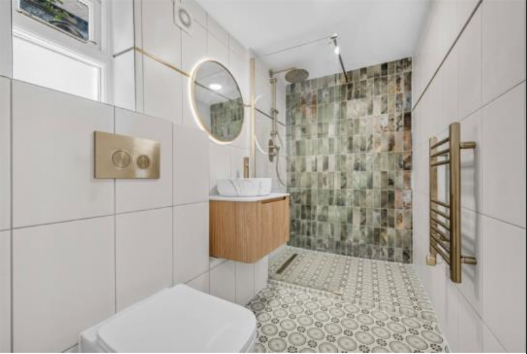 Modern bathroom with white tiled walls and patterned floor, wooden vanity with vessel sink, round mirror, small window, and walk-in shower with textured tile wall and wooden towel rack.