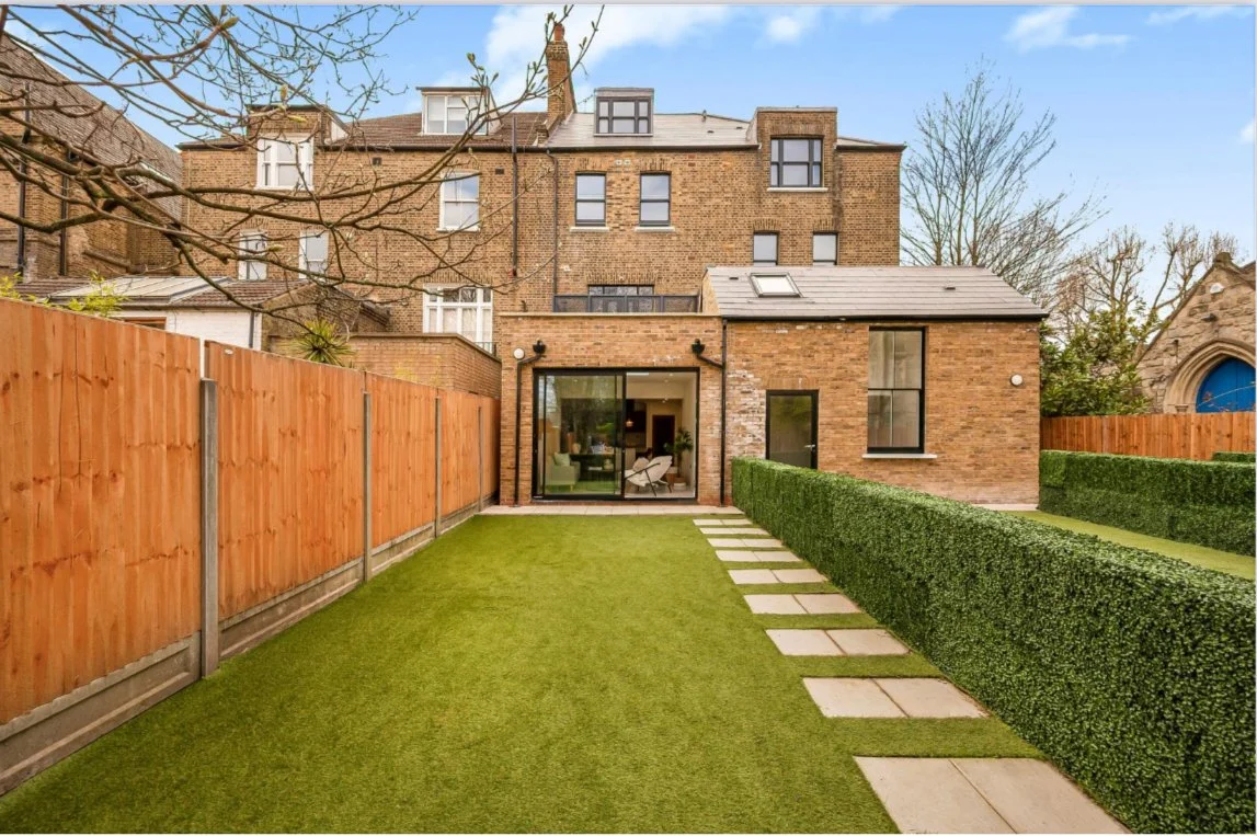 Backyard with artificial grass, stone pathway, and trimmed hedge, view of a brick house with patio doors and windows.