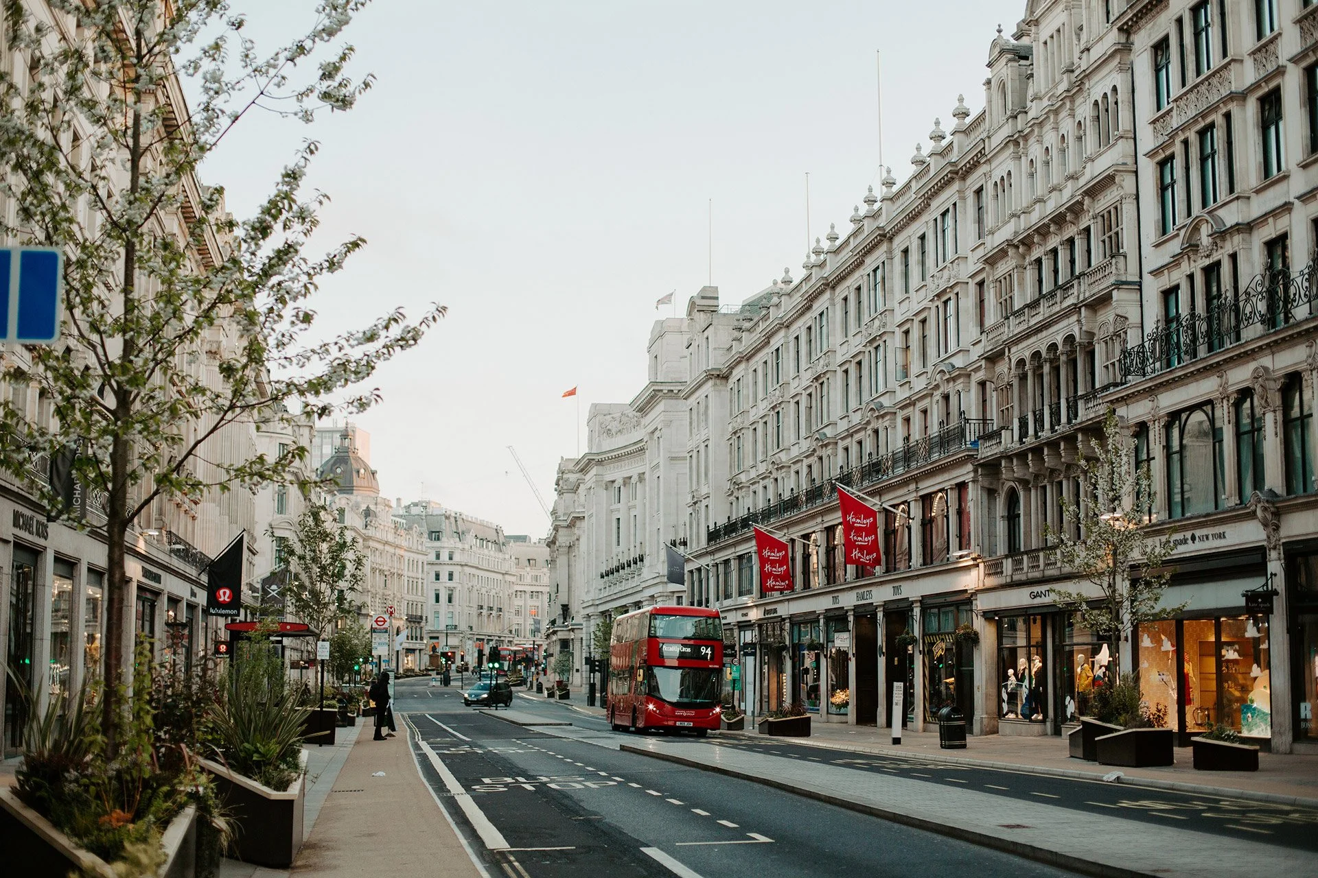 A street scene in London with white historic buildings, a red double-decker bus, and storefronts with red signs.