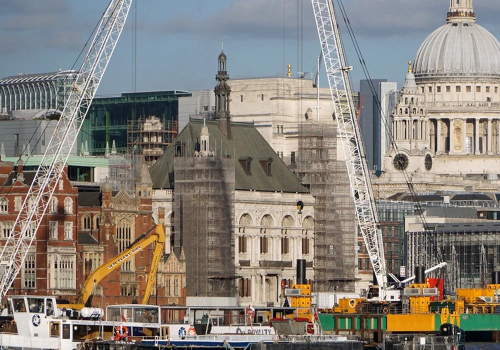 Construction site on a waterfront with cranes, scaffolding, historic and modern buildings, and St. Paul's Cathedral in the background in London.