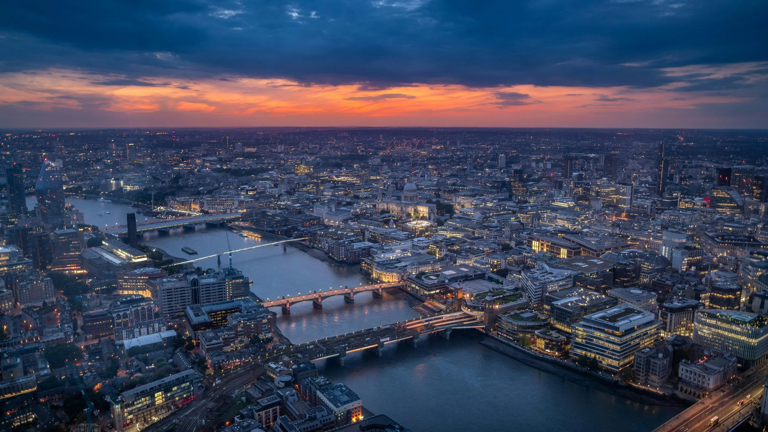 Aerial view of London's skyline at sunset, showing the River Thames, bridges, and illuminated buildings.