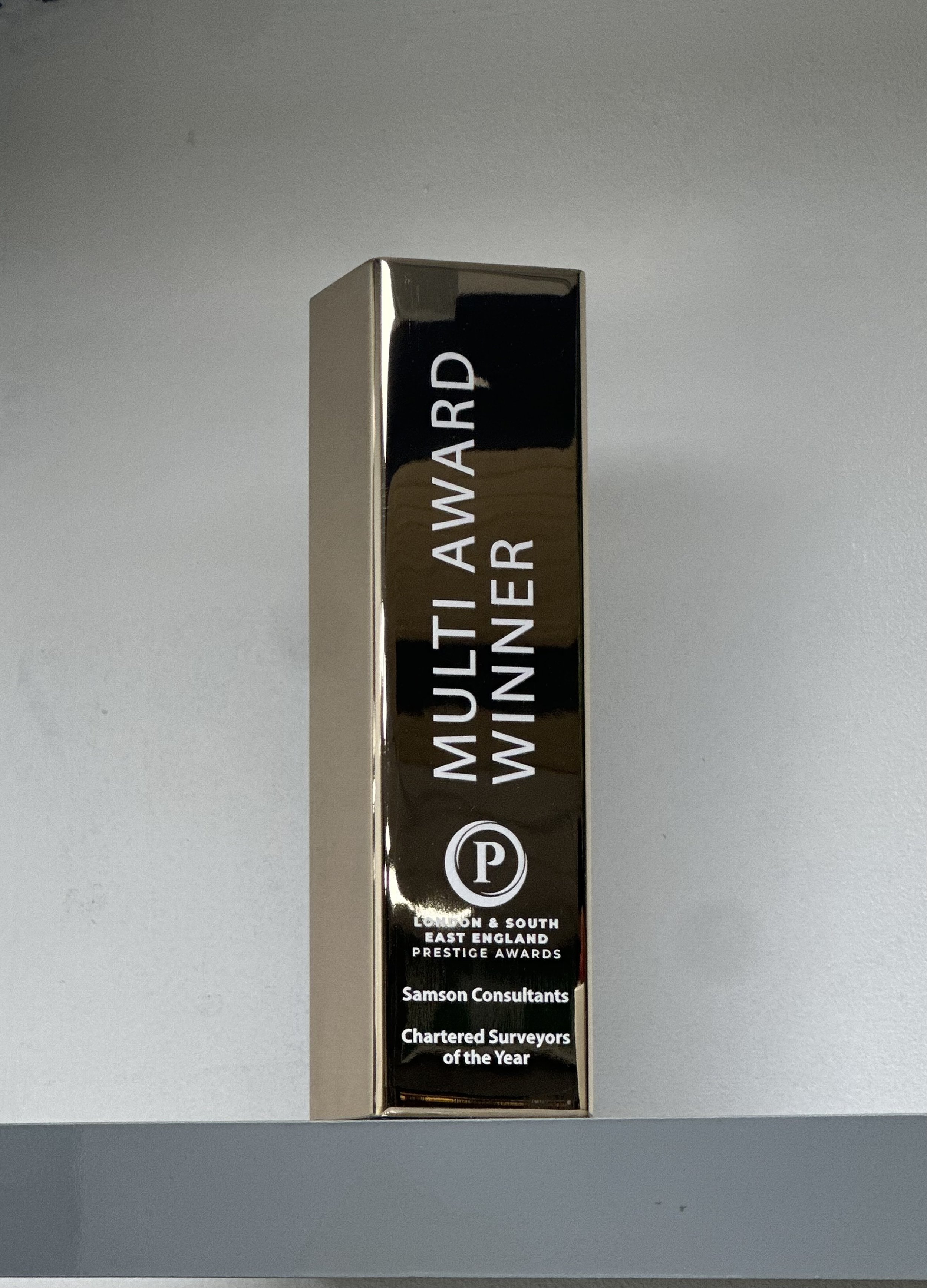A tall, shiny, metallic award trophy with the inscription 'Multi Award Winner' and a logo with the letter 'P'. The text also mentions 'London & South East England Prestige Awards', 'Samson Consultants', and 'Chartered Surveyors of the Year'.