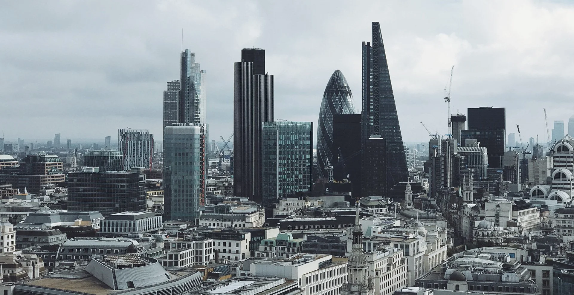 A city skyline with modern high-rise buildings and cranes under a cloudy sky.