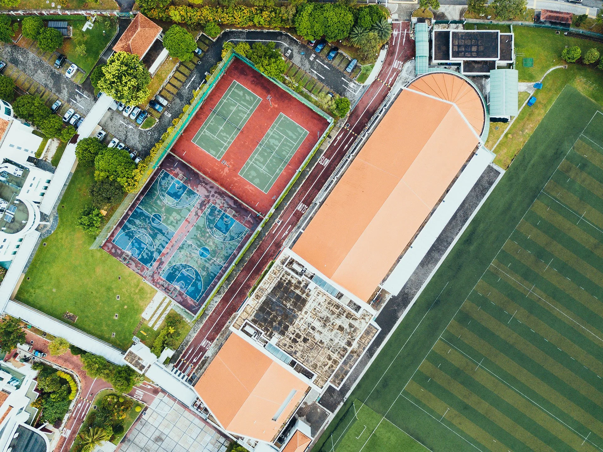 An aerial view of a sports complex showing two basketball courts, two tennis courts, a soccer field, and a parking lot surrounded by trees and buildings.