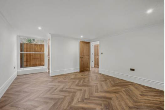 Empty room with white walls, hardwood herringbone flooring, open window, and wooden doors.
