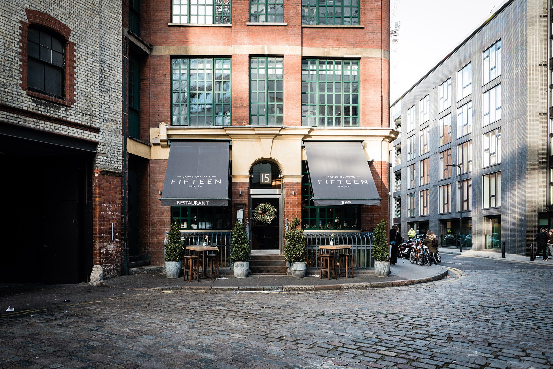 Exterior view of a restaurant and bar called 'Fifteen' located on a street corner in an urban area. The building has brick and stone elements with green window frames. Two awnings above the entrance display the restaurant's name, with outdoor seating including small tables and potted plants in front.