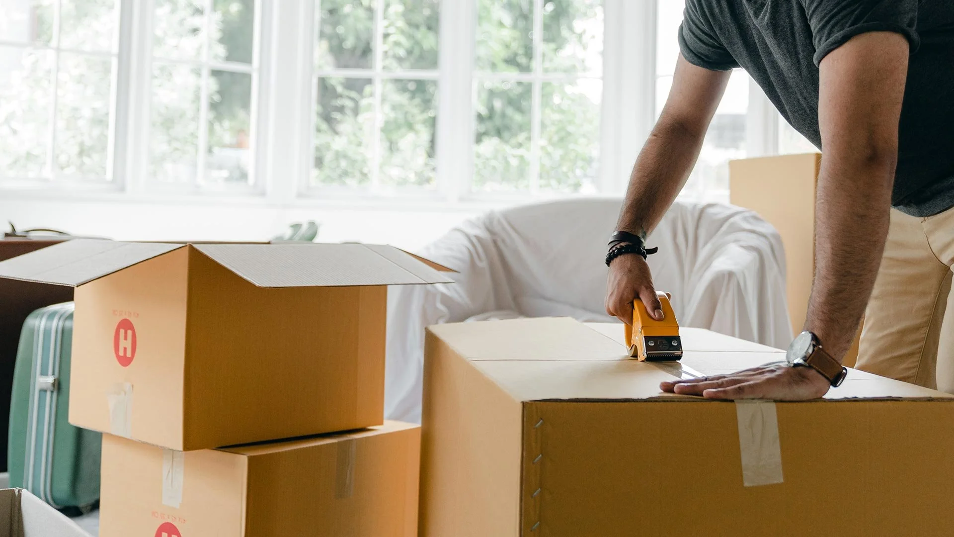 Person using a box cutter to cut a cardboard box inside a bright, sunny room with large windows.