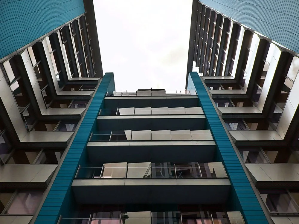View of a modern high-rise apartment building taken from the ground looking up, showing multiple balconies and windows with a partly cloudy sky above.