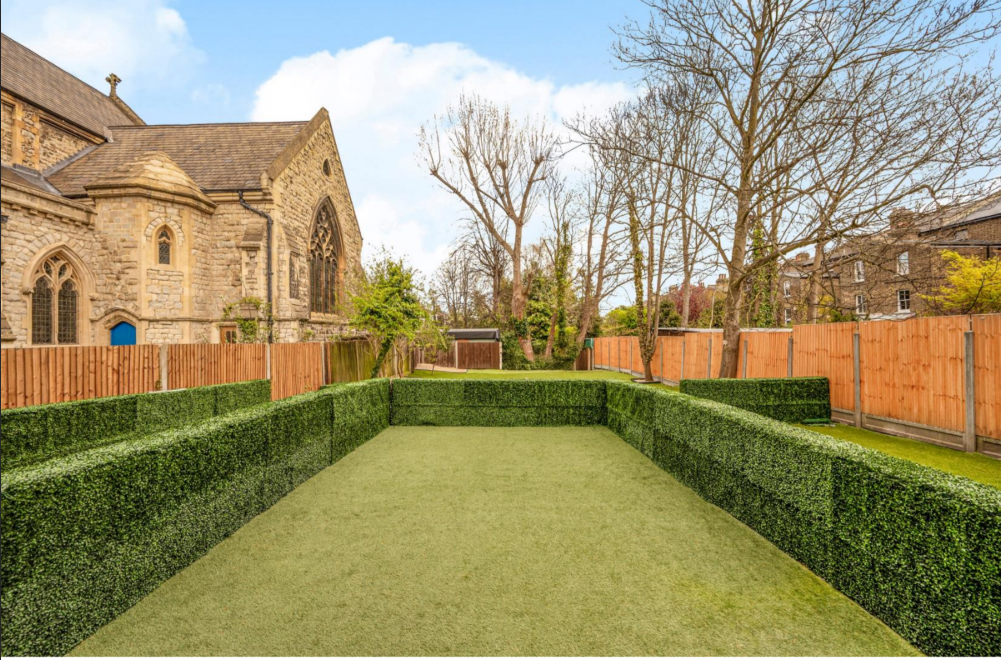A backyard with a rectangular area enclosed by trimmed hedge fences, artificial turf ground, wooden fence, neighboring house, large trees without leaves, and an old stone church on the left.
