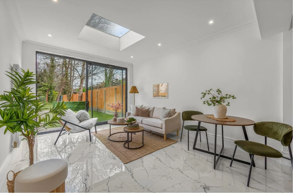 Modern living room with white walls, marble floor, a large window with sliding glass door leading to a backyard, a white sofa, a gray armchair, a wooden dining table with green chairs, and a large potted plant.