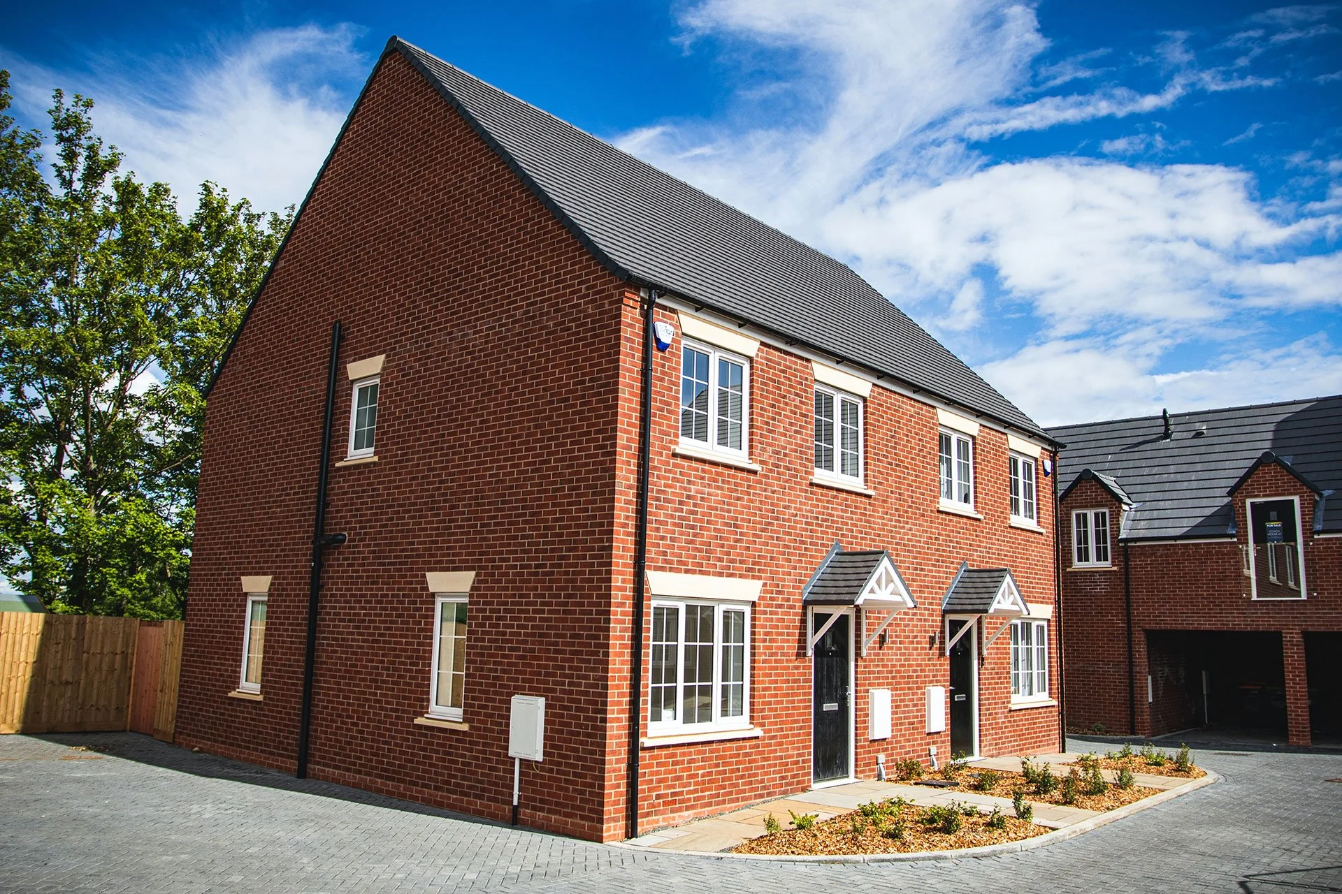 A modern red brick house with white window frames, black doors, and a gray shingled roof, situated on a paved driveway with small landscaped garden beds in front, under a blue sky with wispy white clouds.