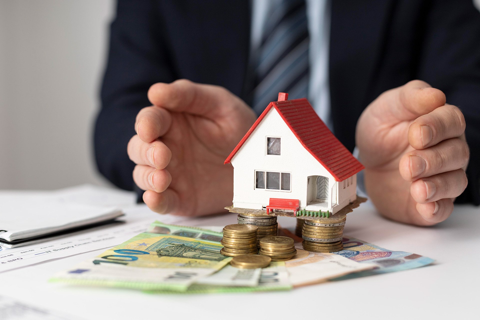 Man in business suit placing a small model house on stacks of coins, with euro banknotes and a pen on a table.