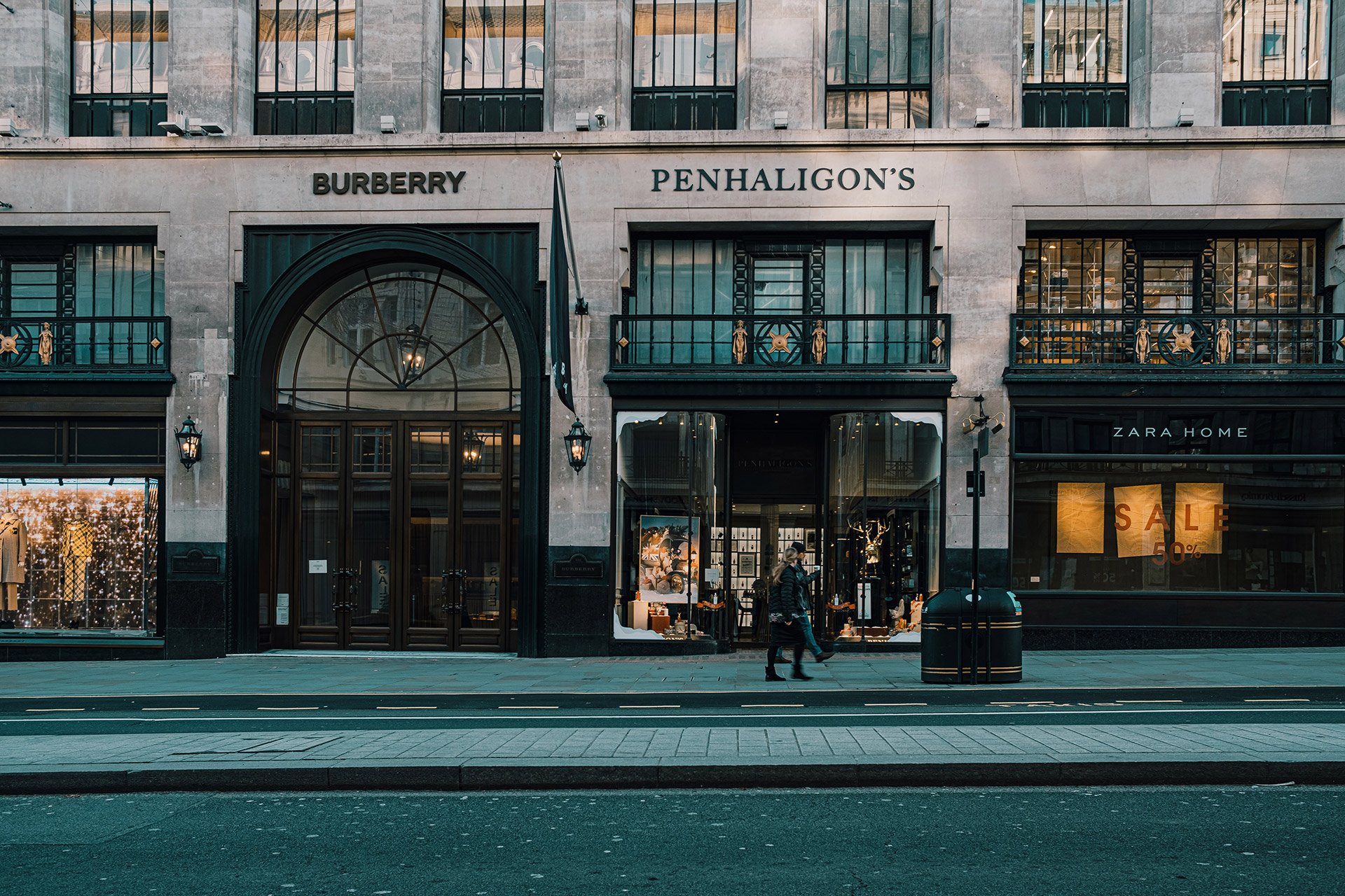 Exterior of a high-end retail store with signs for Burberry, Penhaligon's, and Zara Home, featuring large display windows, a central glass door, and a pedestrian walking by on the sidewalk.