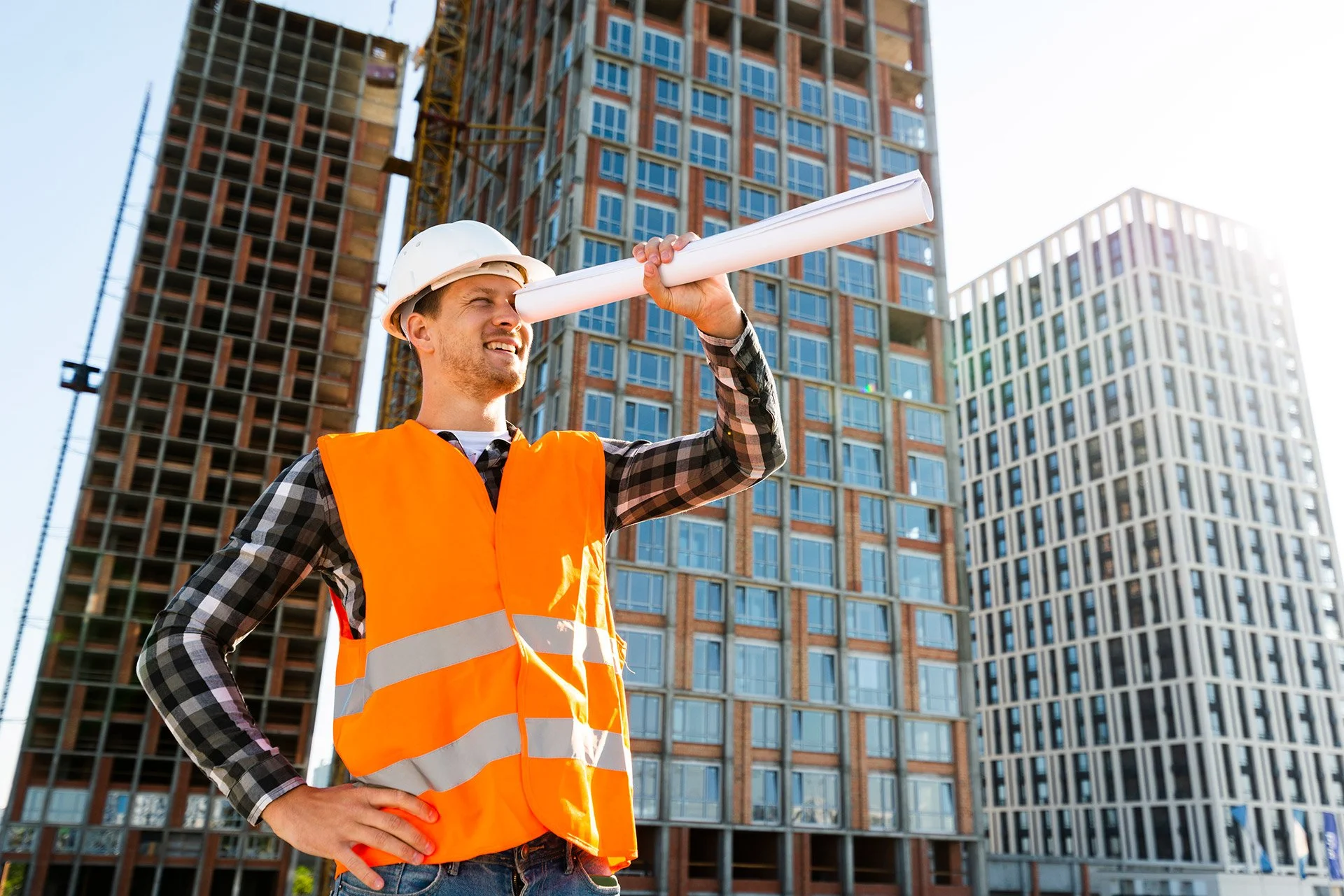 Construction worker in safety vest and helmet holding rolled-up blueprint in front of tall modern buildings under construction.