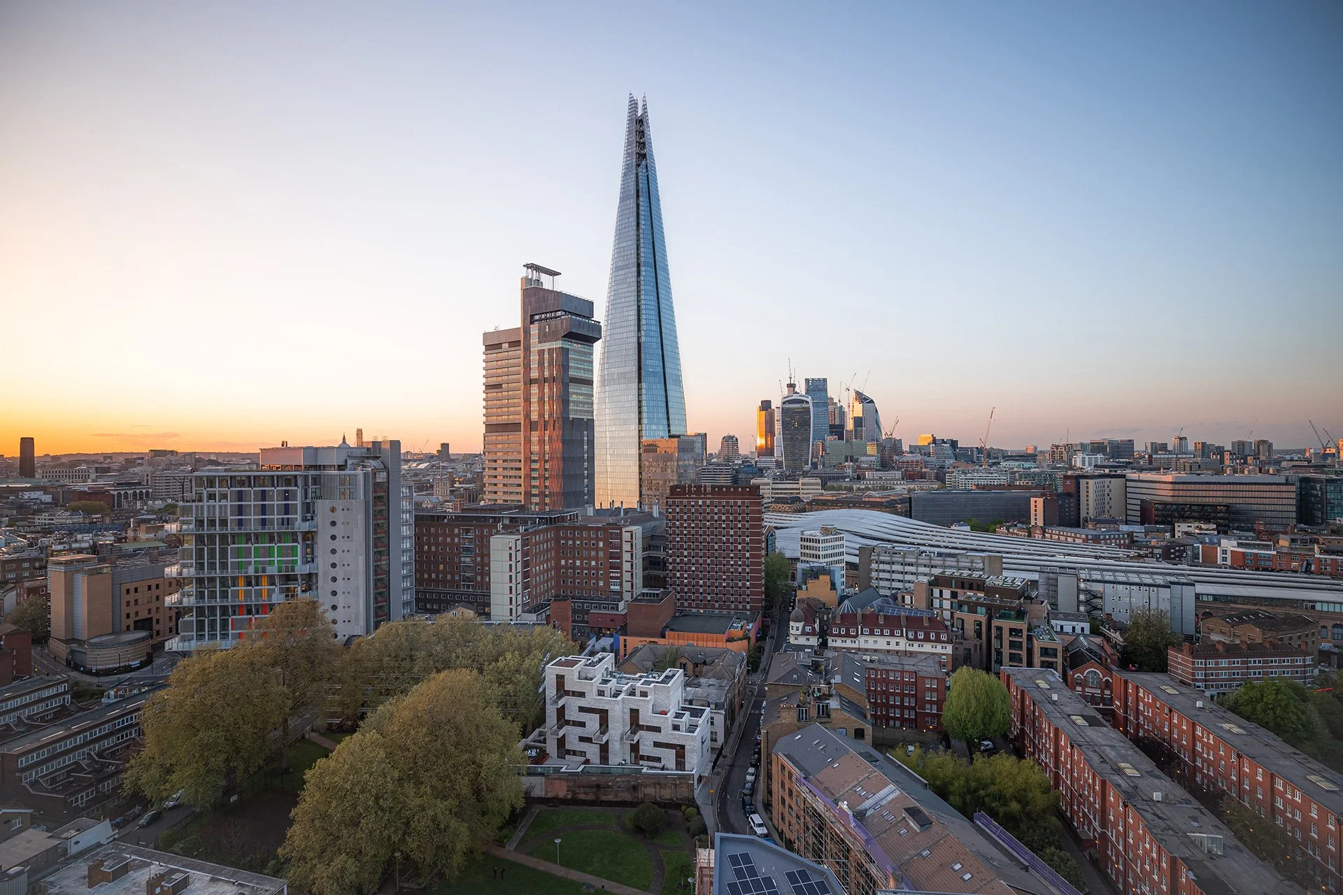 City skyline featuring the Shard skyscraper during sunset with modern buildings and green trees in foreground.