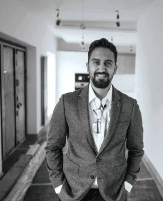 A man in a blazer smiling, standing in an interior hallway.