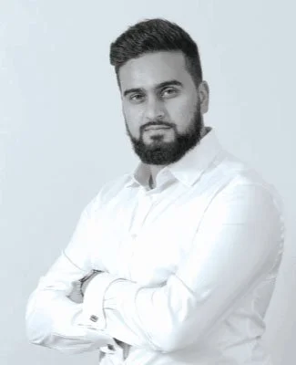 Black and white portrait of a man with a beard and styled hair, wearing a white button-up shirt, with arms crossed.