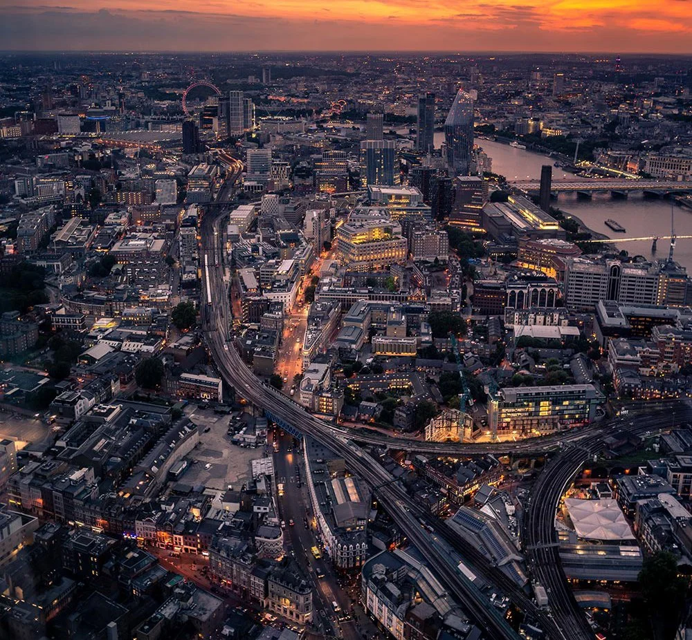 Aerial view of a city skyline at sunset with high-rise buildings, a river running through the city, and a network of roads and bridges illuminated by streetlights.